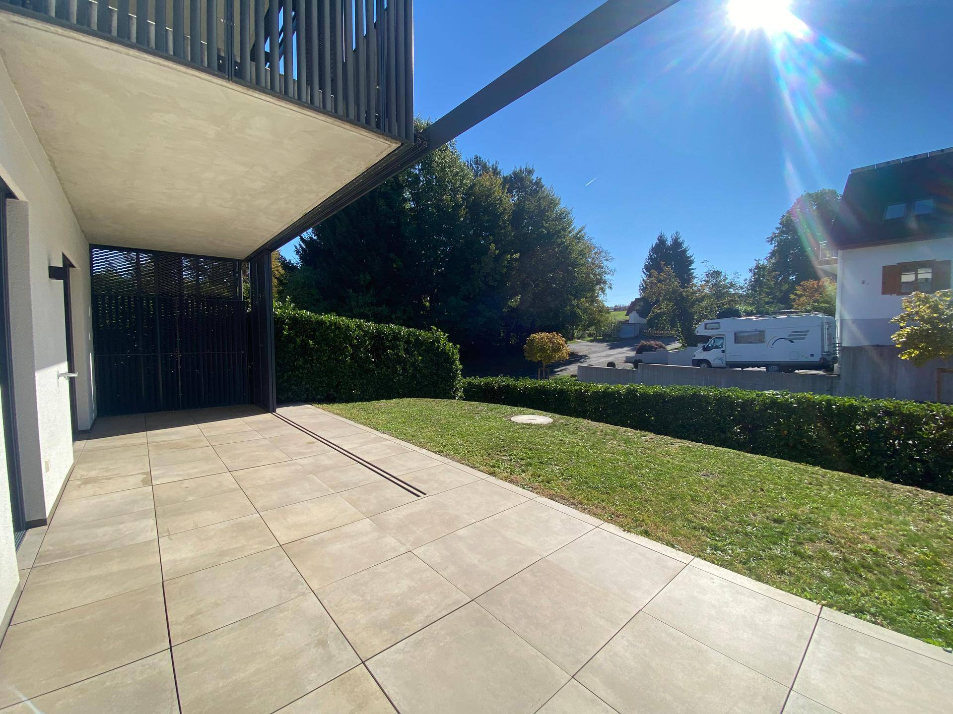 Sonniger Terrassenbereich mit Fliesenboden und angrenzendem Rasen, umgeben von grünen Hecken.