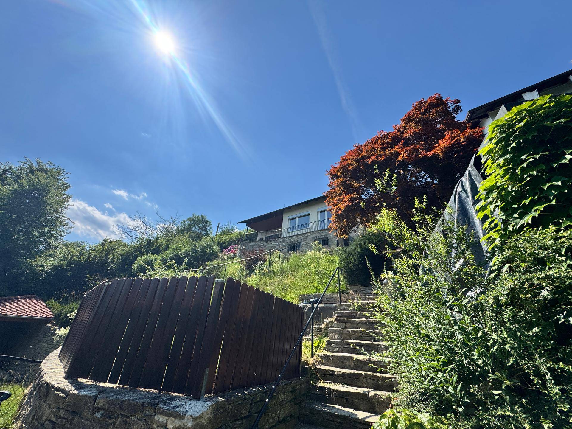 Steintreppe und Mauerwerk führen zu einem Haus auf einem bewachsenen Hang unter strahlendem Sonnenschein.