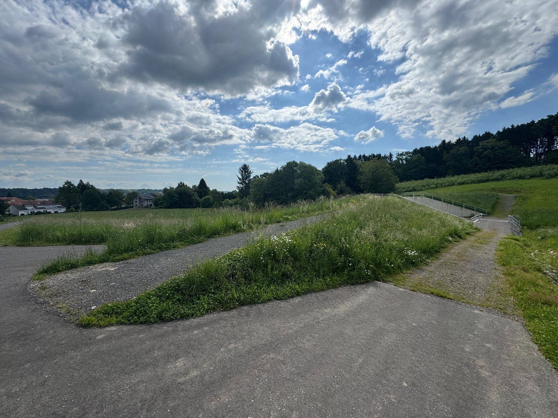 Geteilter asphaltierter Weg durch eine grüne Landschaft mit Hügeln und Betonstrukturen im Hintergrund.