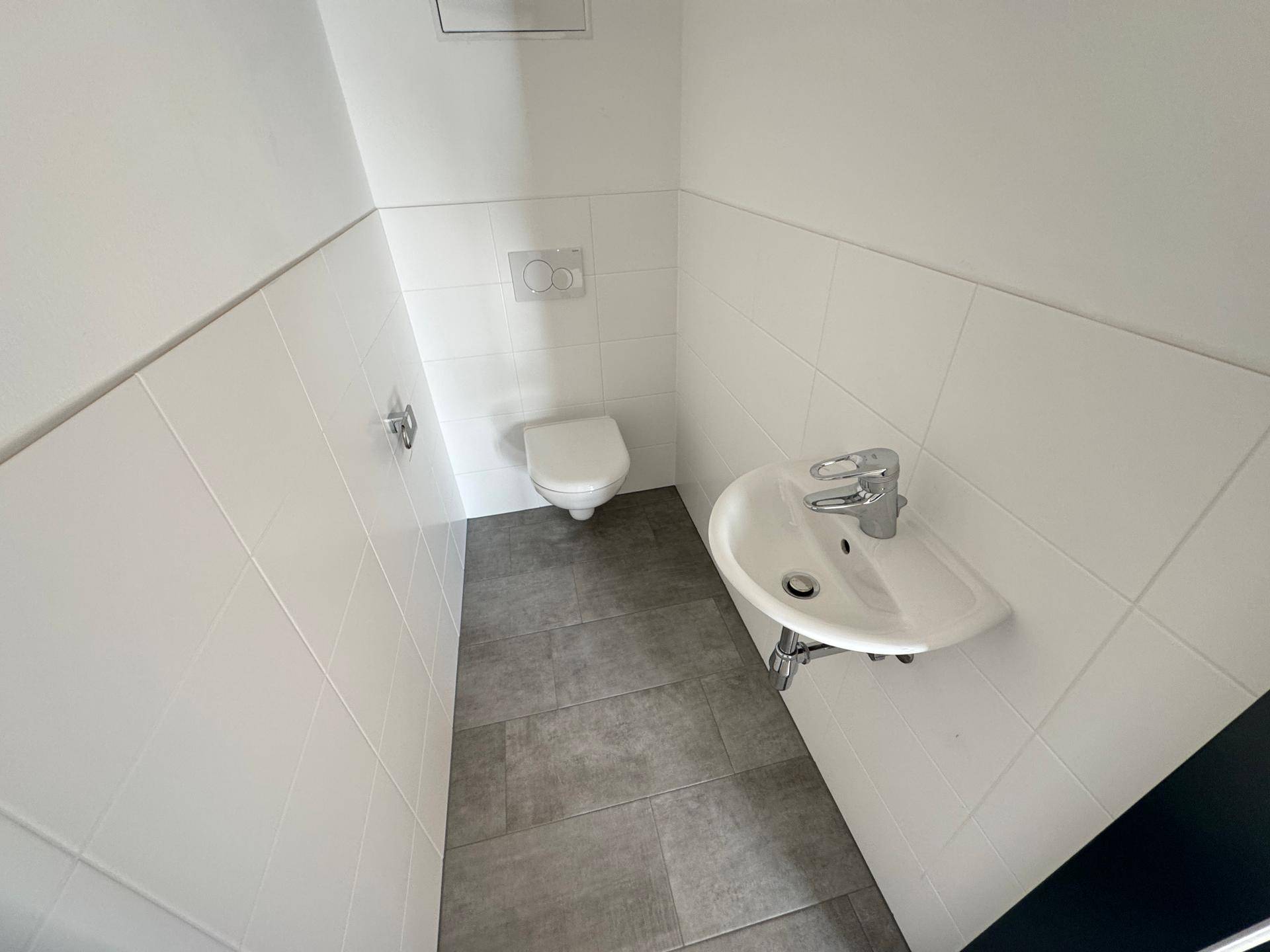 Modernes Gäste-WC mit Wand-WC, kleinem Waschbecken und hellen Fliesen.
