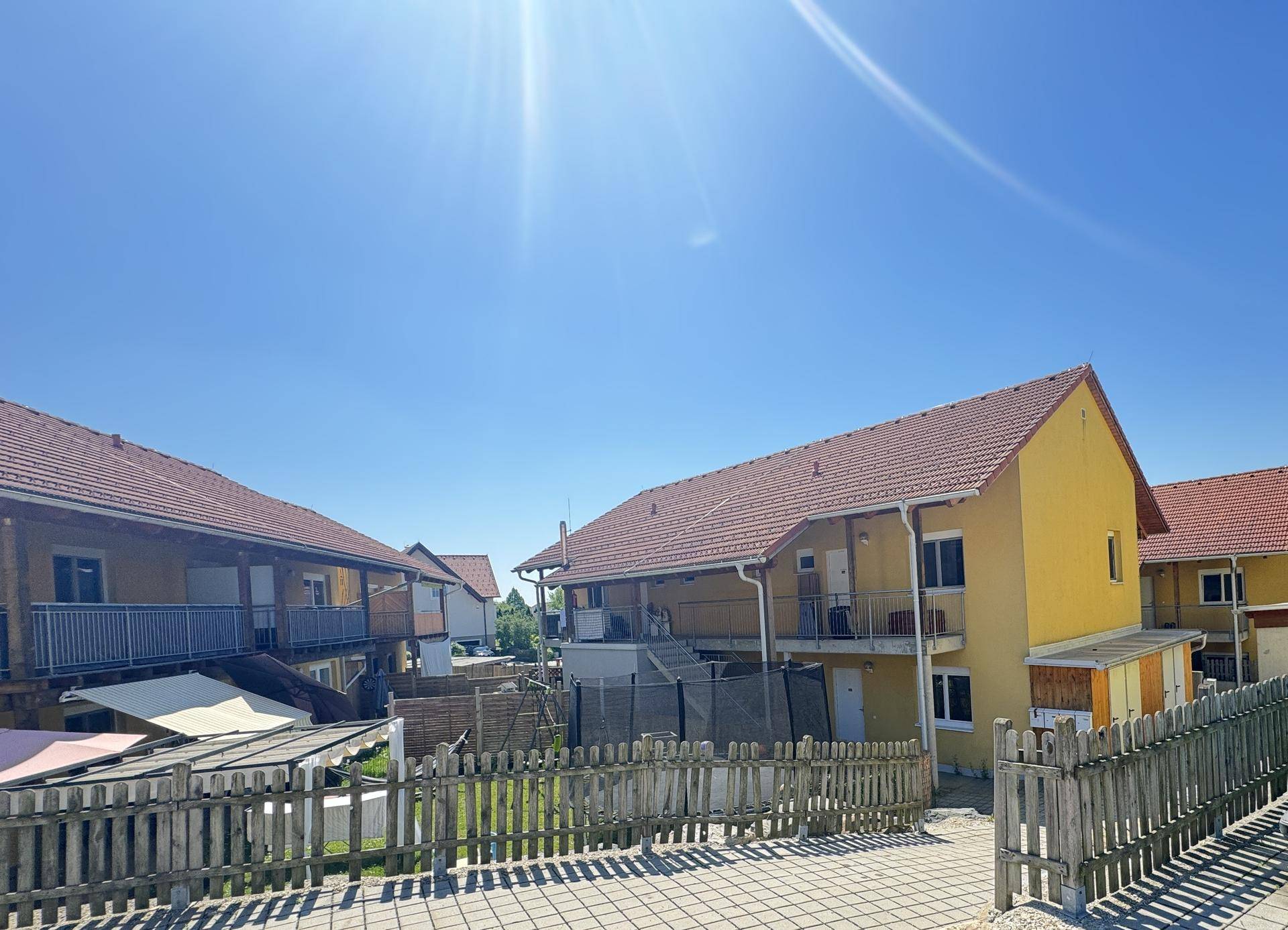 Mehrere Wohngebäude mit Holzzäunen und roten Dächern unter strahlendem Sonnenschein.