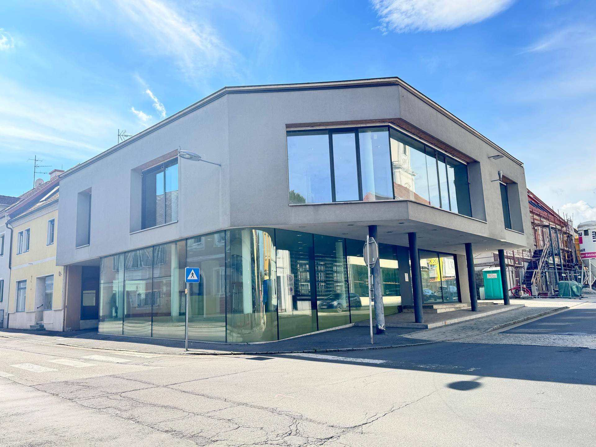 Zeitgenössisches Gebäude mit großen Fensterfronten und moderner Fassade an einer belebten Straße unter blauem Himmel.