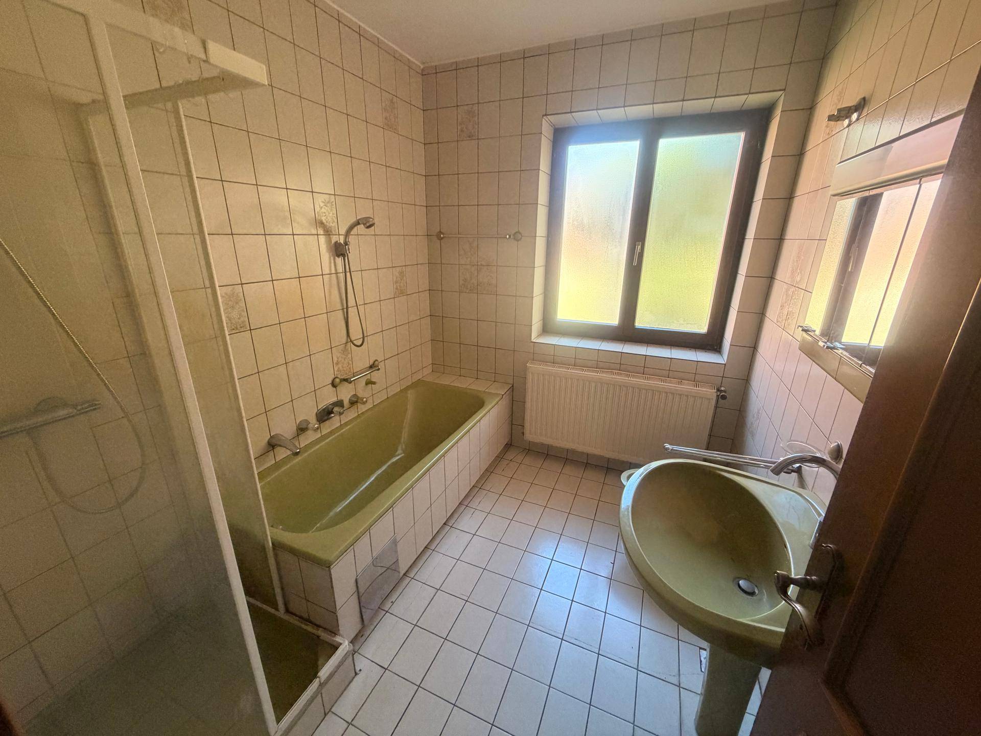 Badezimmer mit grüner Badewanne, Dusche, Waschbecken und Fenster im 80er-Jahre-Stil.
