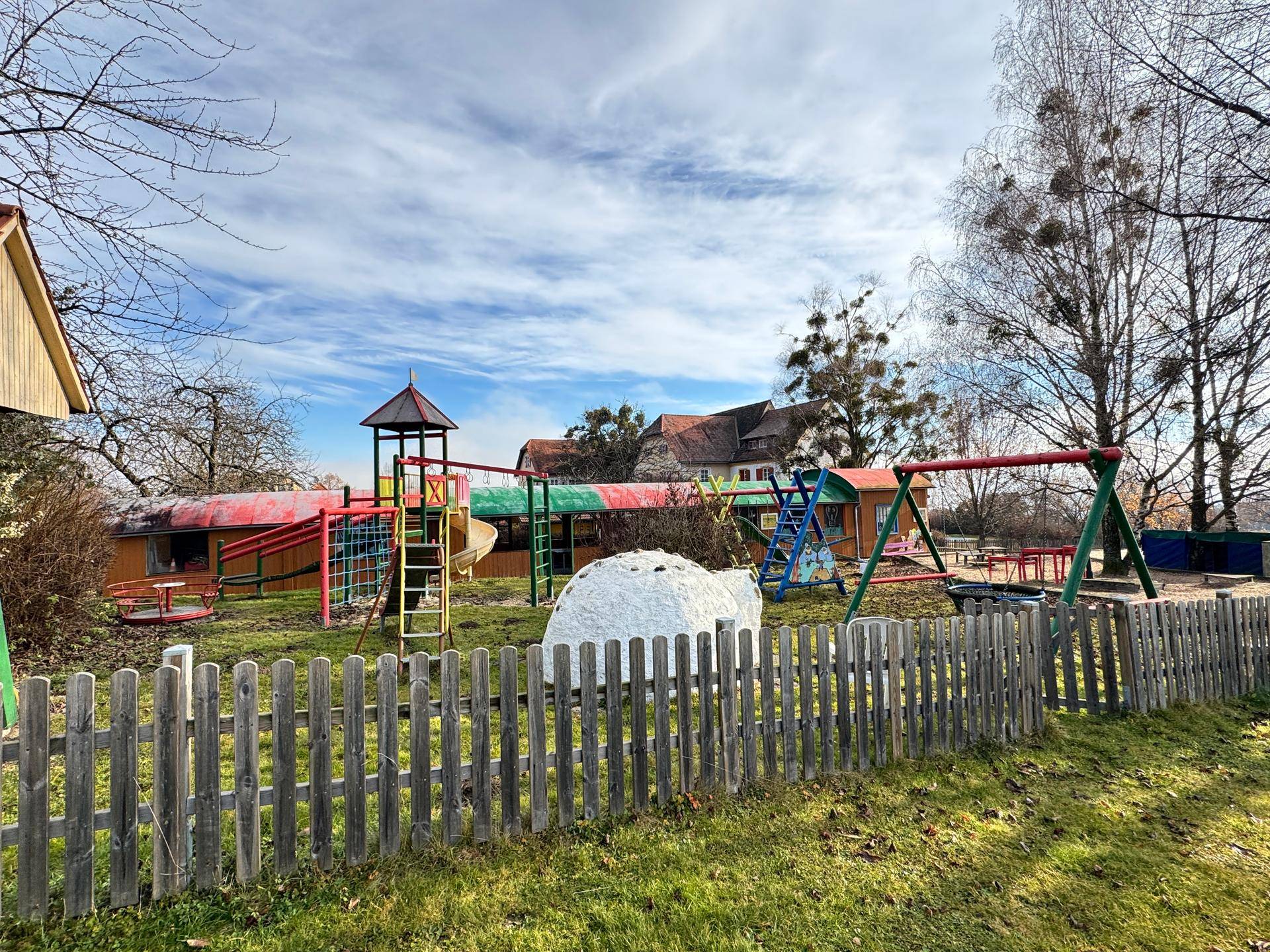 Spielplatz mit verschiedenen Spielgeräten und Holzzaun in einem Gartenbereich.