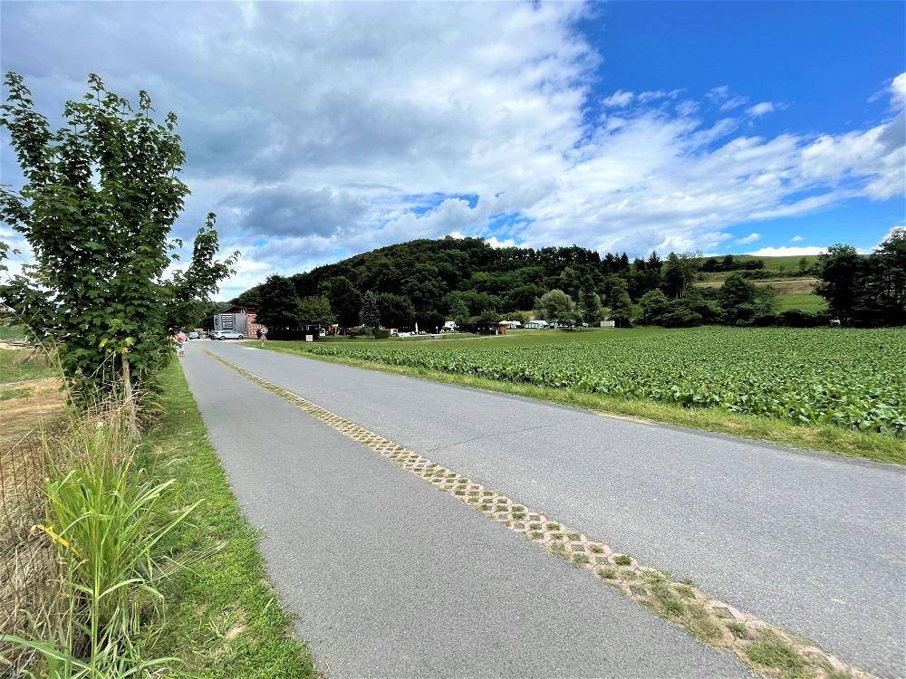 Ländliche Straße mit Feldern und bewaldeten Hügeln unter einem bewölkten Himmel.