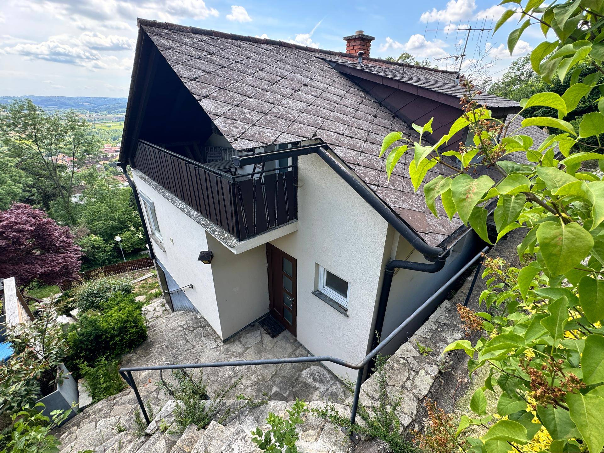 Ansicht des Hauses von oben mit Satteldach, kleinem Balkon und umgebender grüner Hügellandschaft.