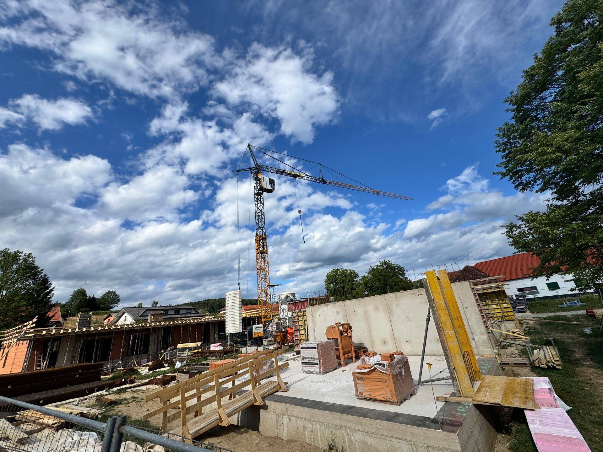 Baustelle mit Betonfundamenten, Holzschalung und einem Baukran vor ländlicher Kulisse.