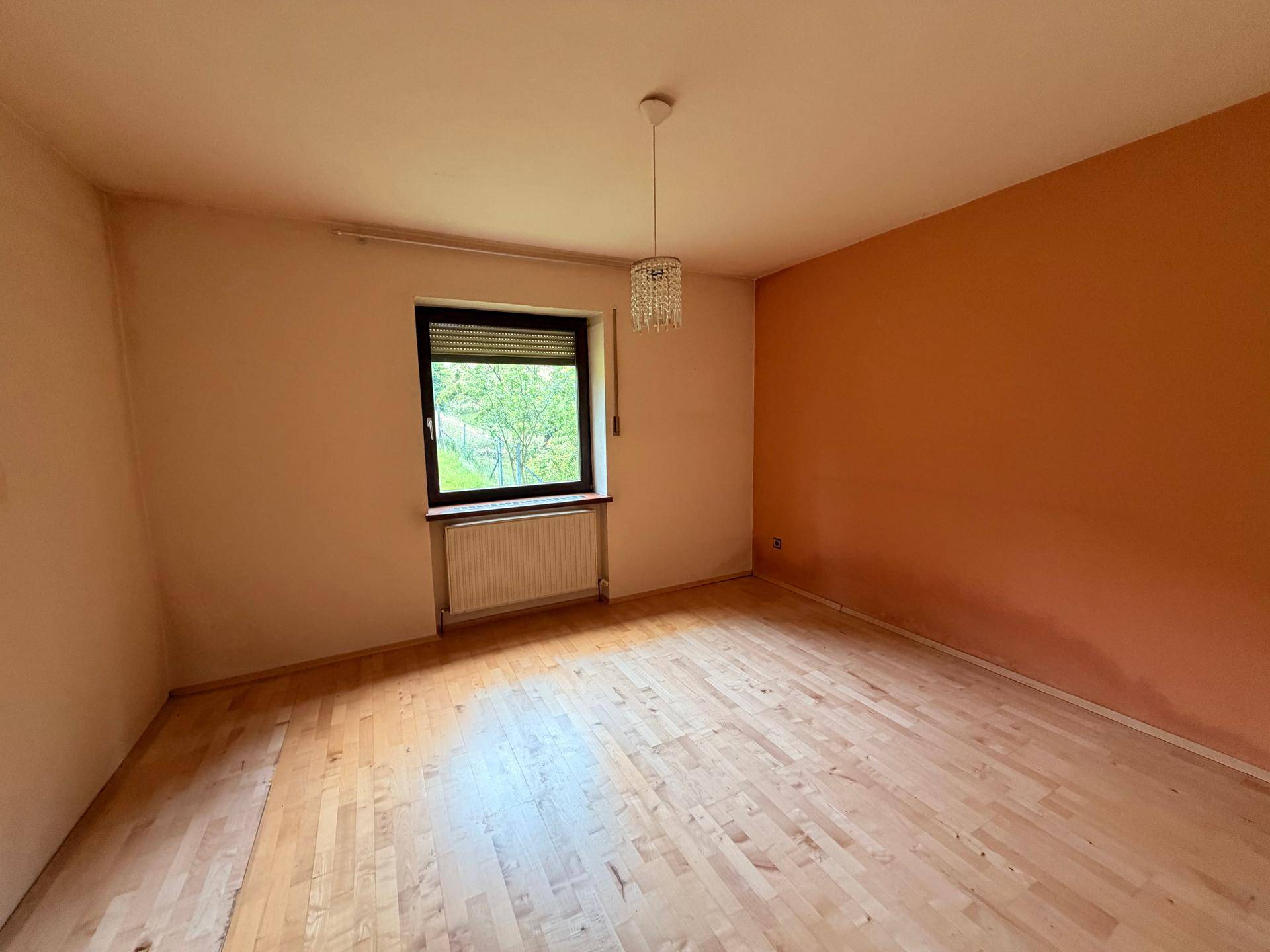 Leeres Schlafzimmer mit Laminatboden, Heizkörper und Fenster mit Rollladen.