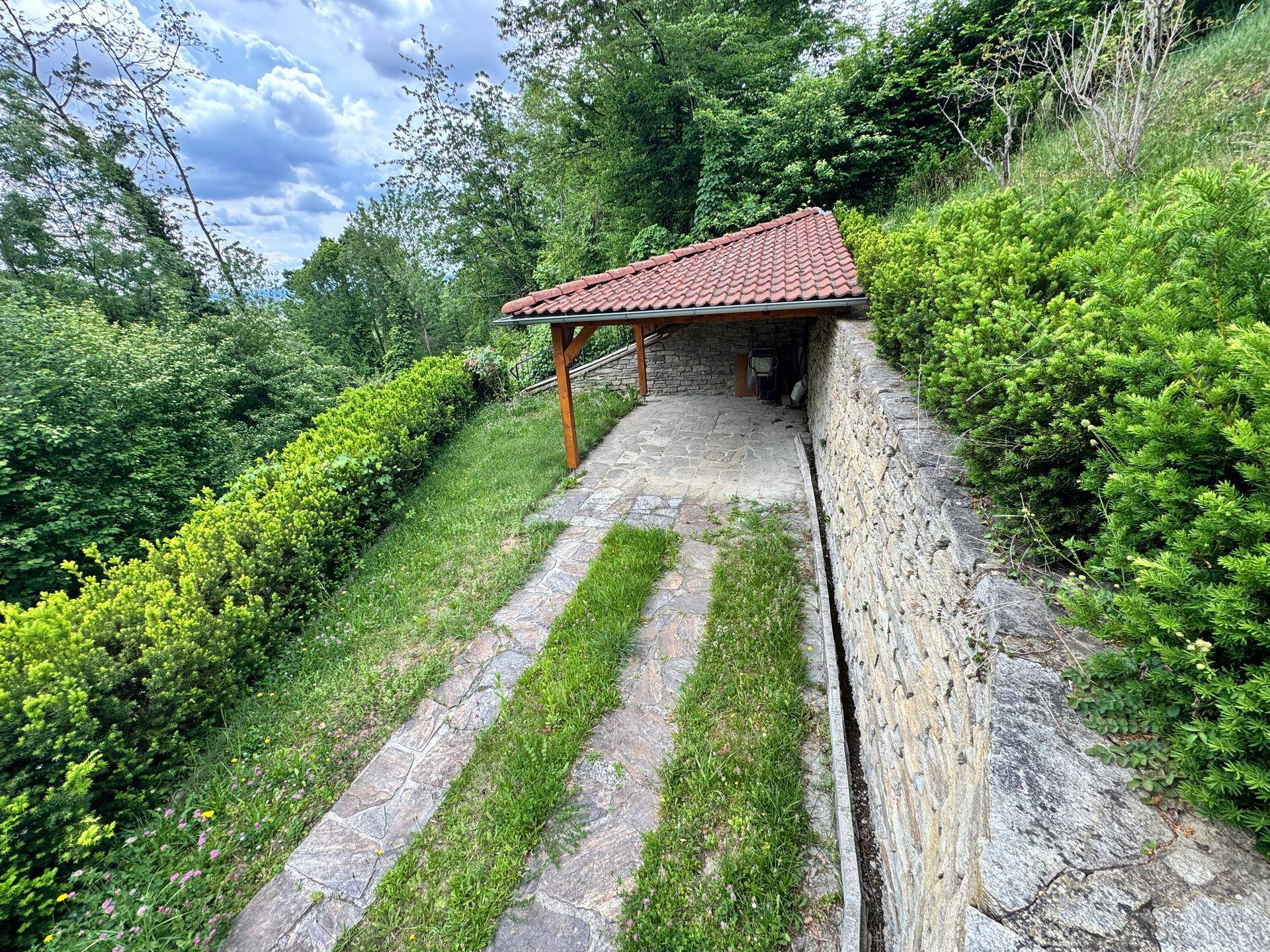 Überdachter Stellplatz oder Schuppen mit Ziegeldach und Steinmauer, umgeben von Hecken.