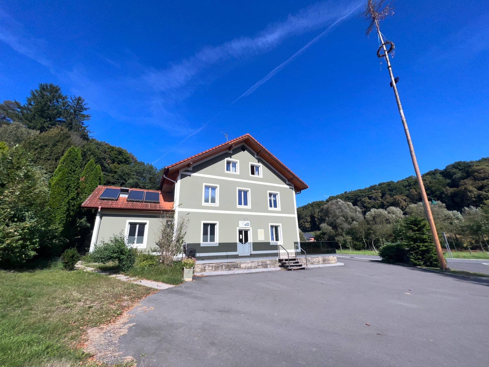 Gebäudeansicht mit Solarpaneelen auf dem Dach, großem Parkplatz und umgebender grüner Landschaft.
