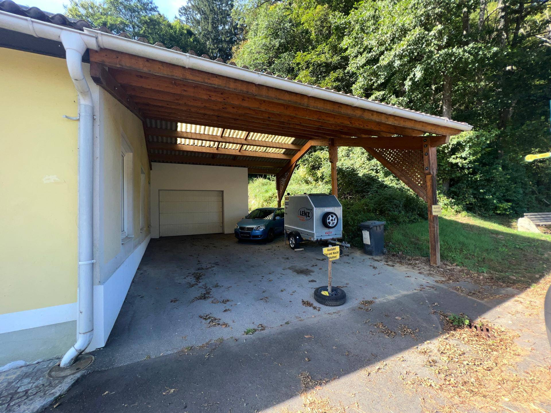 Überdachter Carport-Bereich mit Holzbalken und angrenzender Garage, umgeben von Bäumen.
