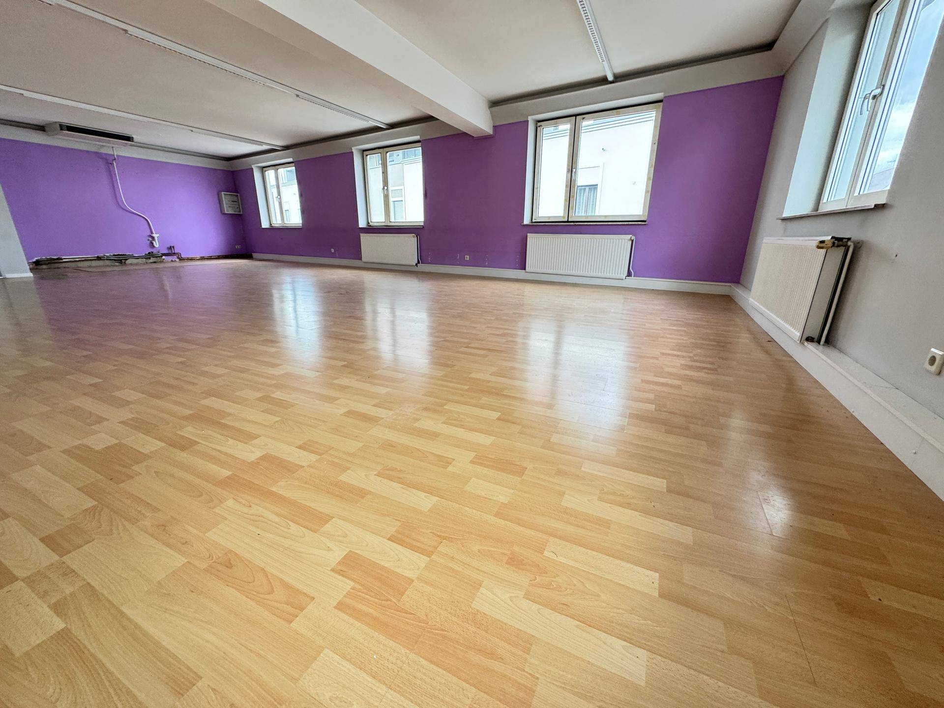 Geräumiges Zimmer mit Laminatboden, Heizkörpern unter den Fenstern und lila Wänden.