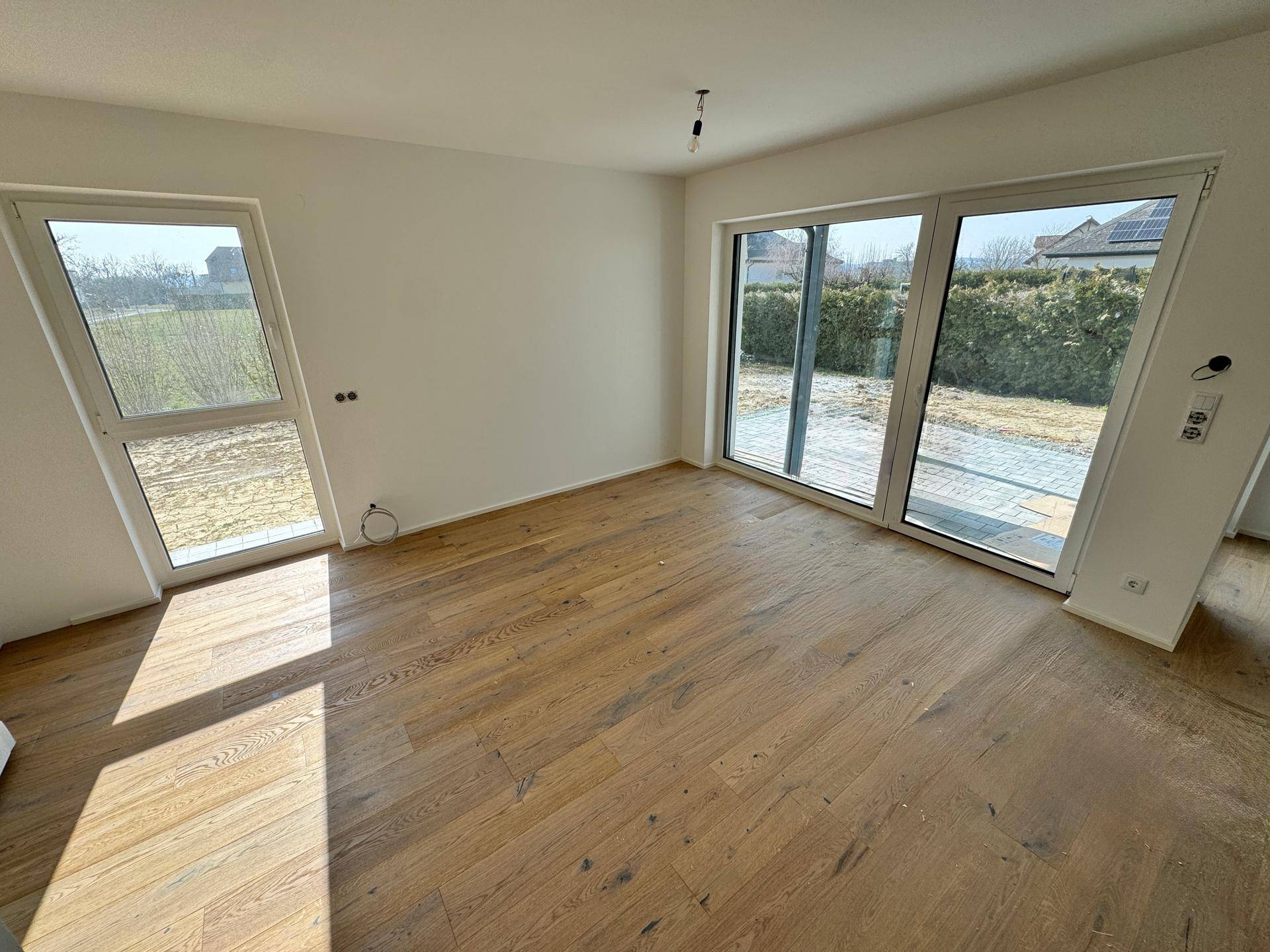 Lichtdurchfluteter Wohnraum mit Holzboden und bodentiefen Fenstern, die Zugang zum Außenbereich bieten.