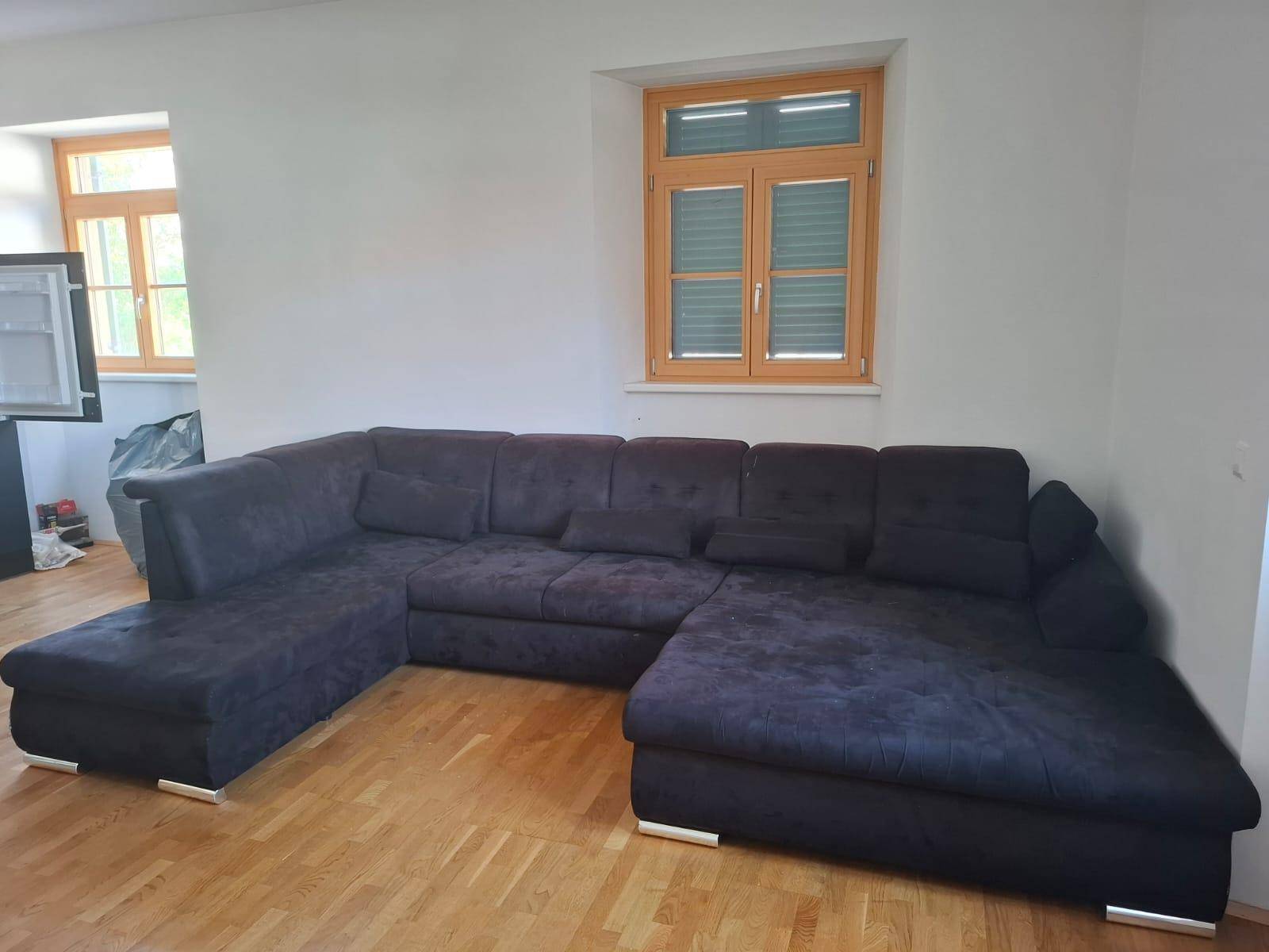 Geräumiges Wohnzimmer mit großem Ecksofa, Holzboden und zwei Fenstern für natürliche Belichtung.