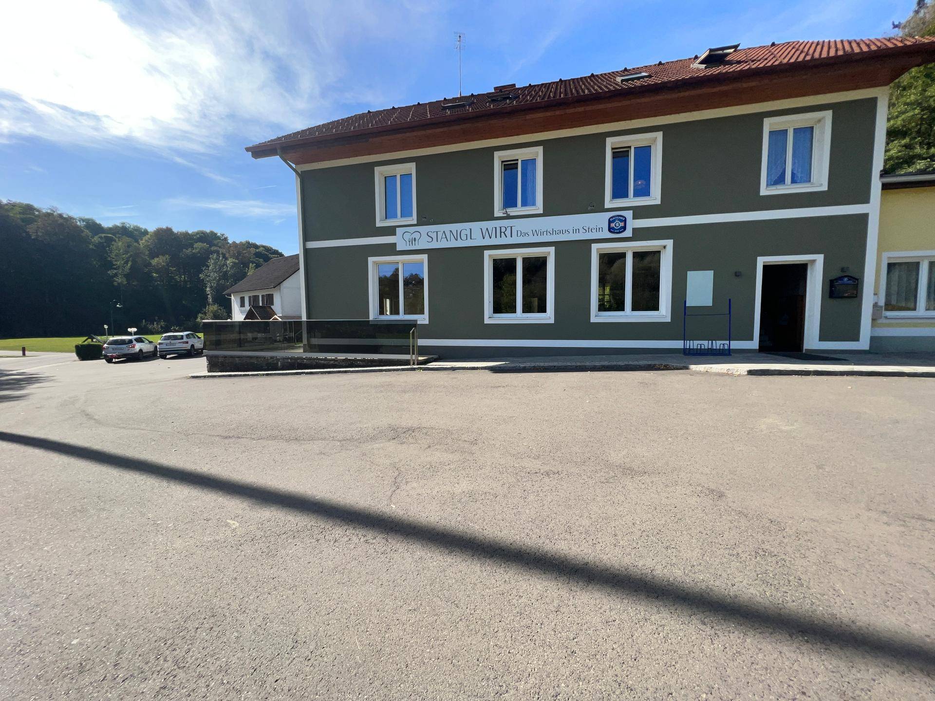 Vorderansicht des Gebäudes mit großem asphaltierten Parkplatz und dem Schriftzug "Stangl Wirt".
