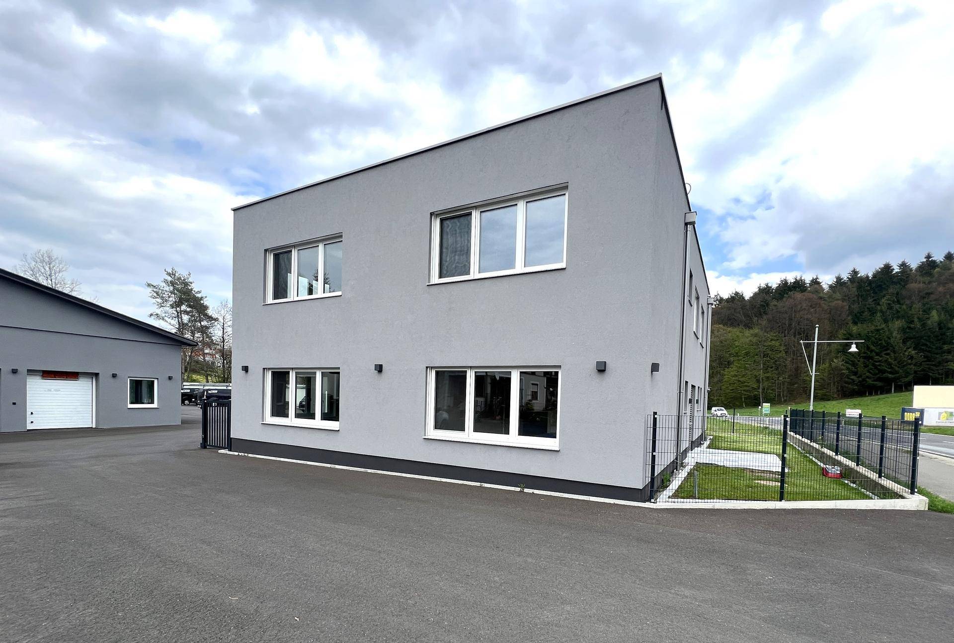Modernes Geschäftsgebäude mit grauer Fassade und asphaltiertem Vorplatz, neben einem weiteren Gebäude gelegen.