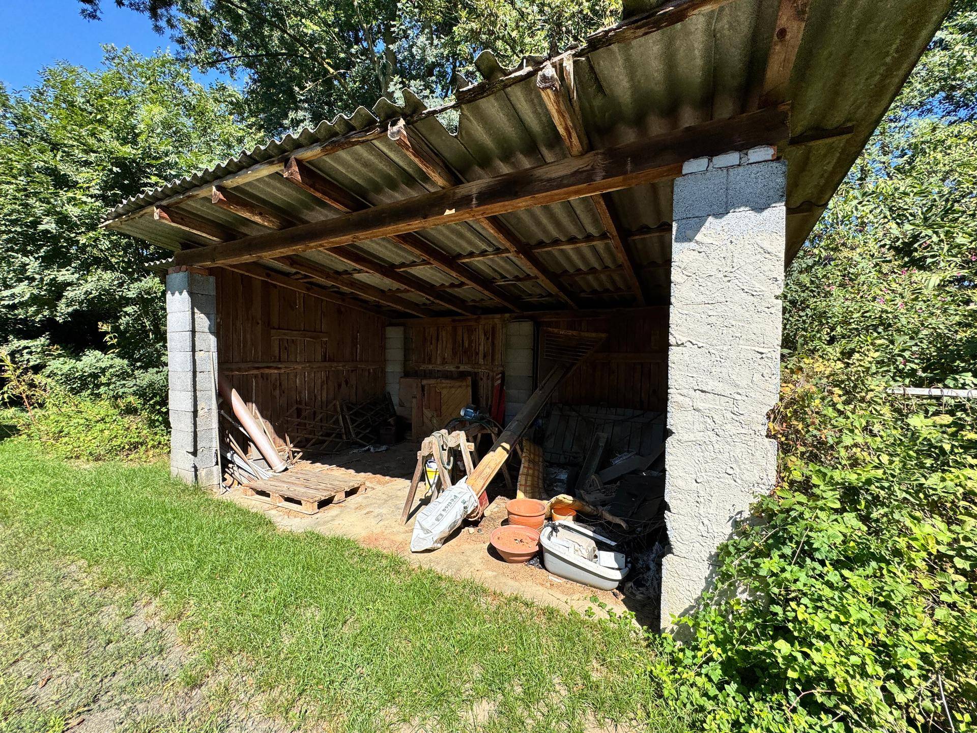 Offener Unterstand im Gartenbereich mit Wellblechdach und verschiedenen Gegenständen.