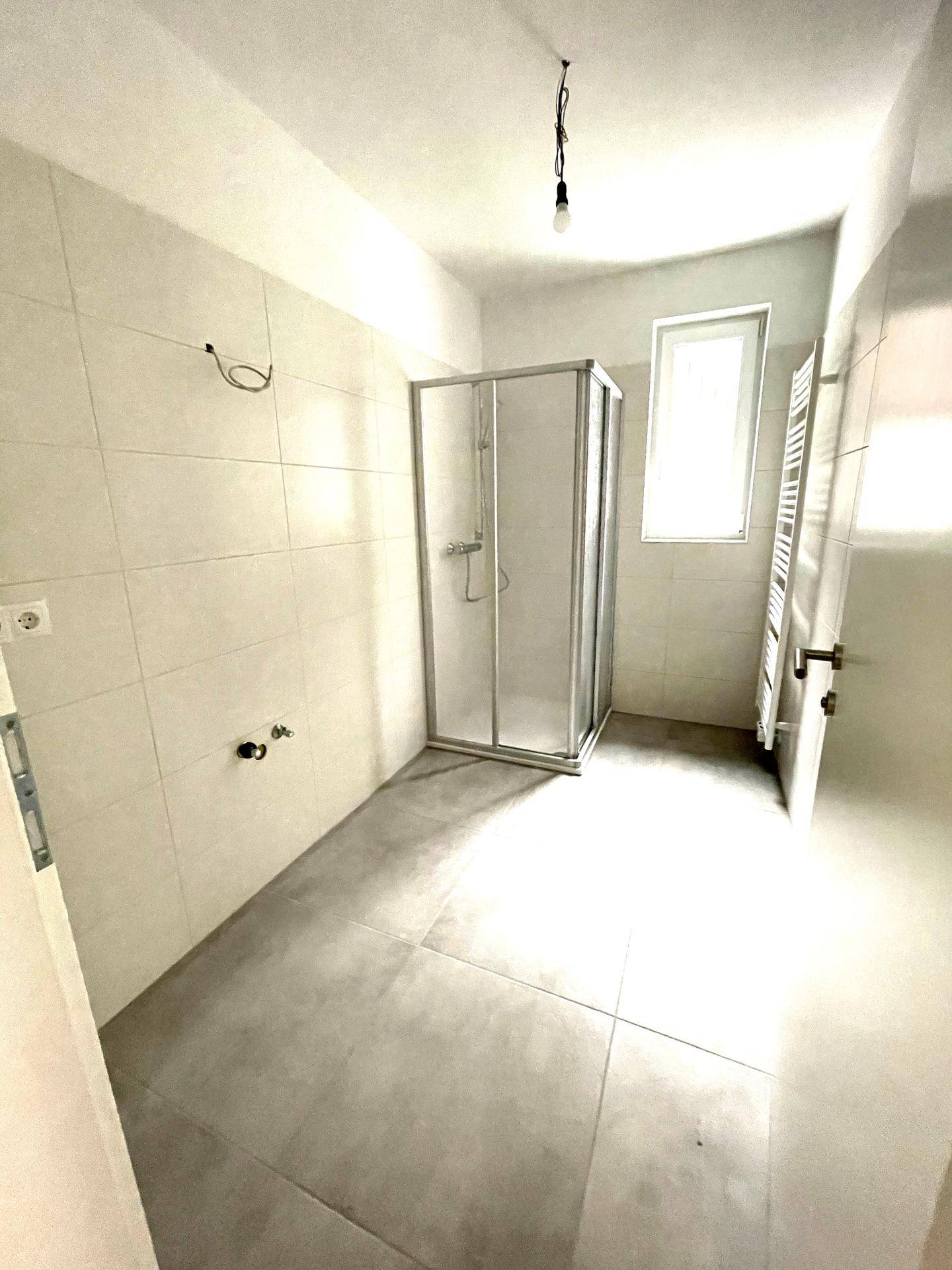 Modernes Badezimmer mit Duschkabine, grauen Fliesen und Fenster für natürliche Belüftung.