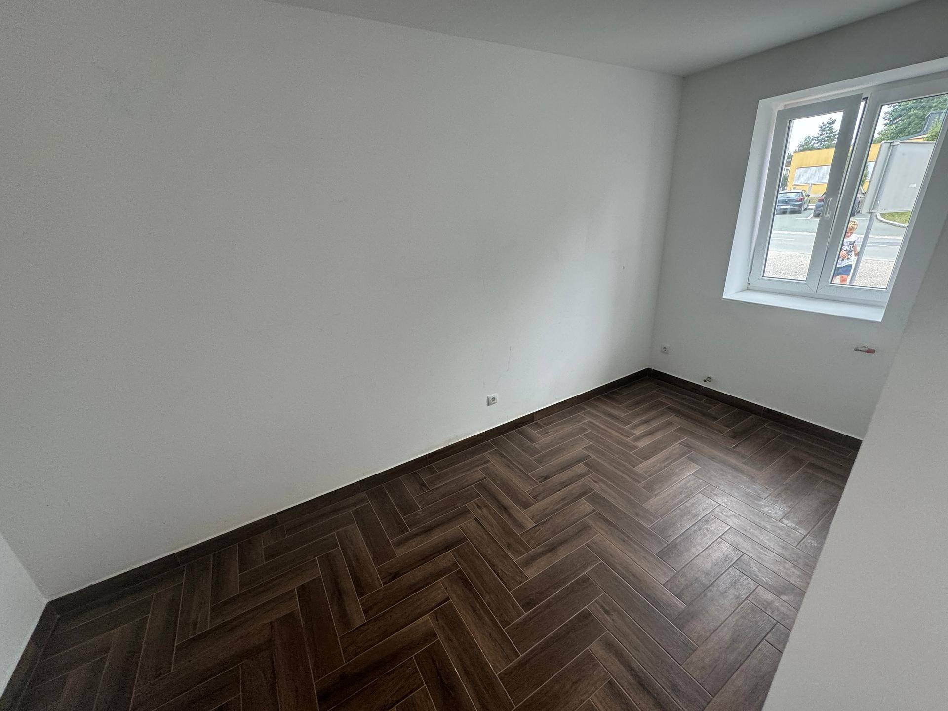 Kleinerer Raum mit dunklem Fischgrätparkett und einem Fenster, das viel Tageslicht hereinlässt.
