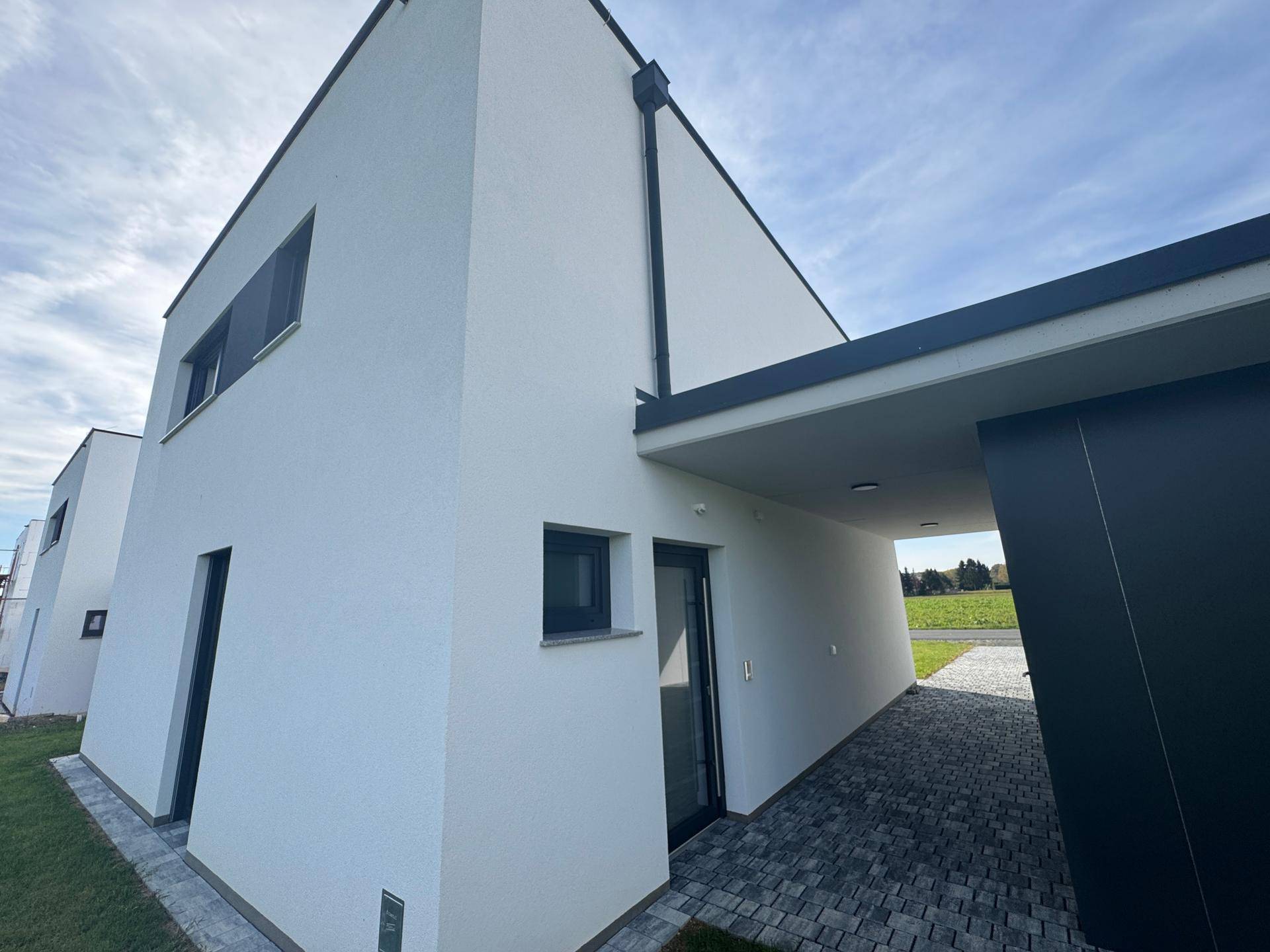 Seitenansicht eines modernen Hauses mit weißer Fassade, Carport und grünen Feldern im Hintergrund.