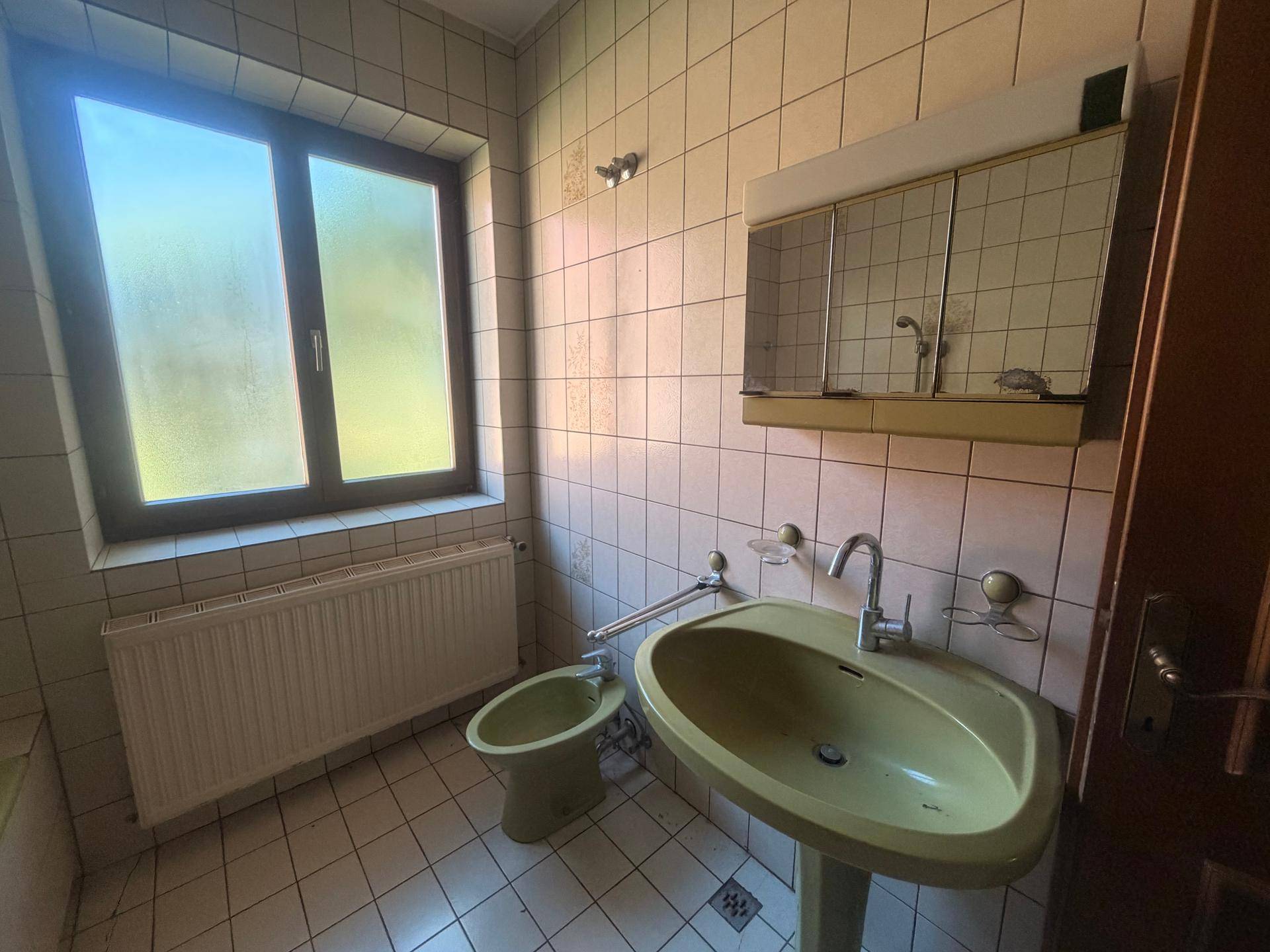 Badezimmer mit grünem Waschbecken, Bidet, Fenster und Spiegelschrank im 80er-Jahre-Stil.