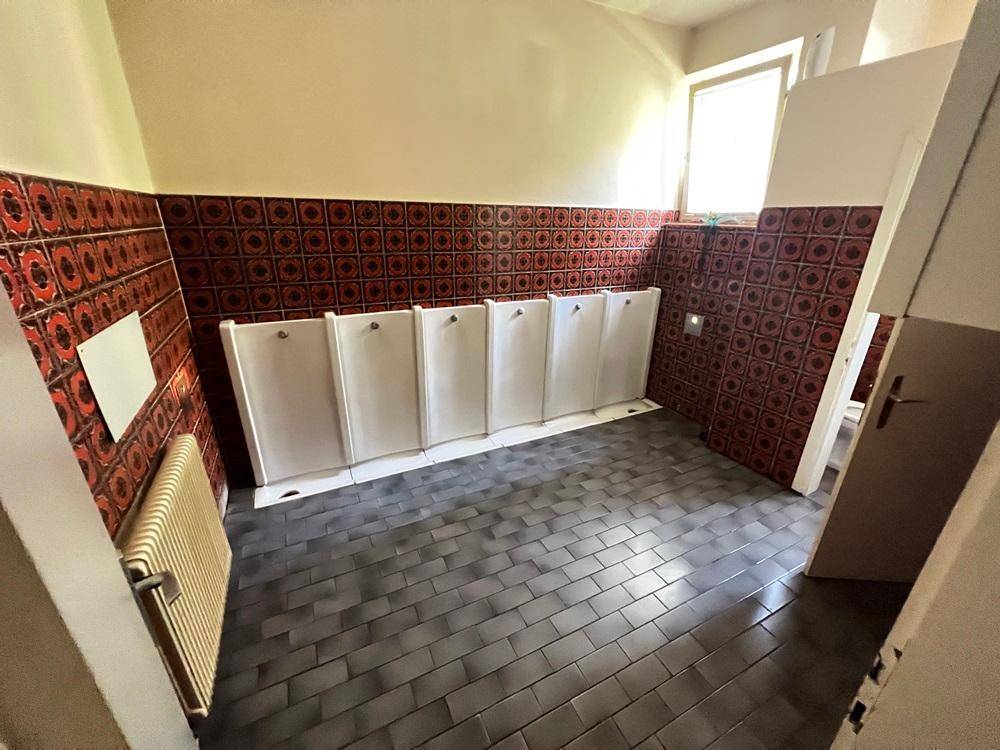 Sanitärraum mit mehreren Urinalen, roten Fliesen und einem Fenster für Tageslicht.