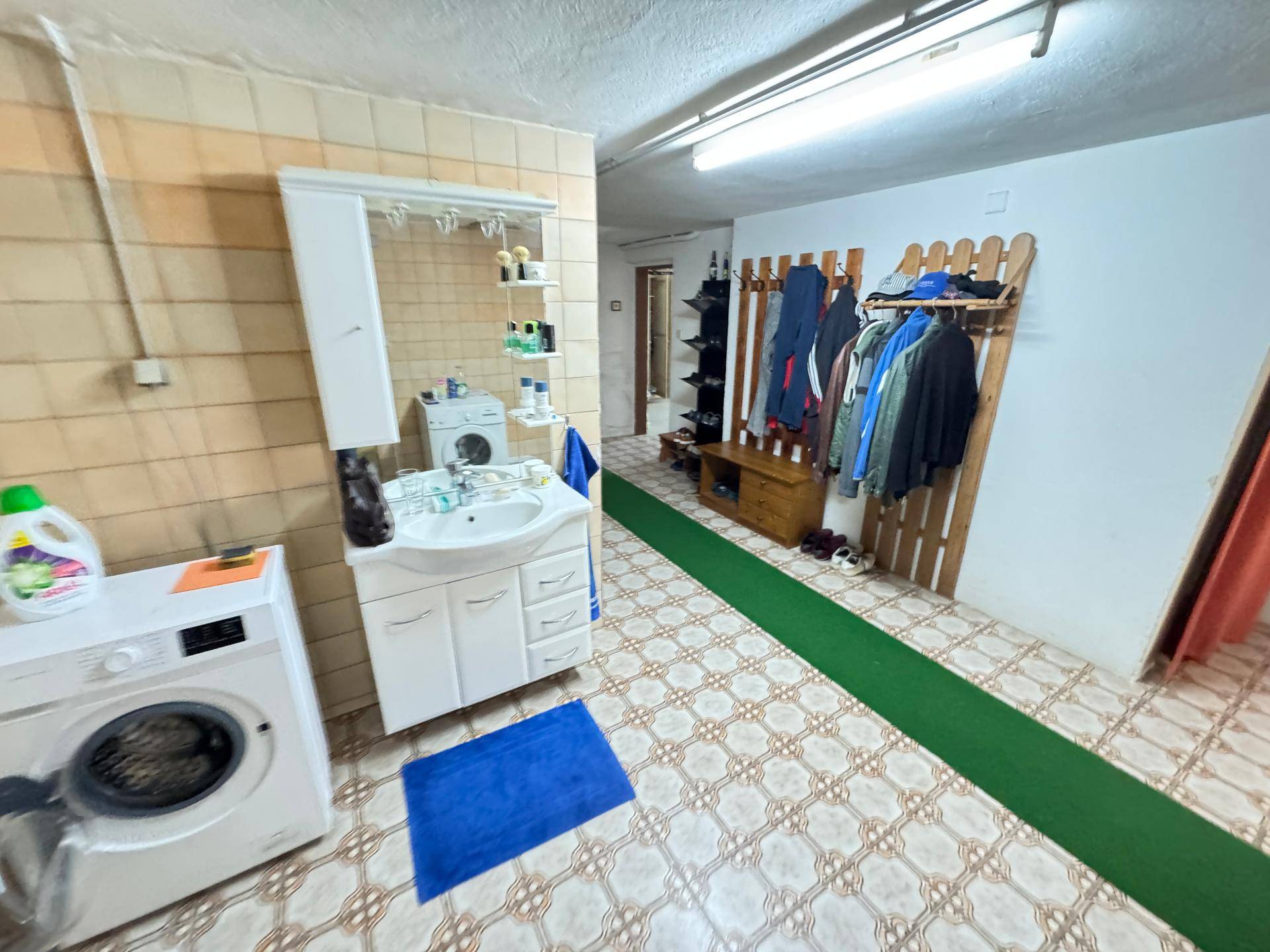 Waschküche im Untergeschoss mit Waschmaschine, Waschbecken und Garderobe, gefliestem Boden.