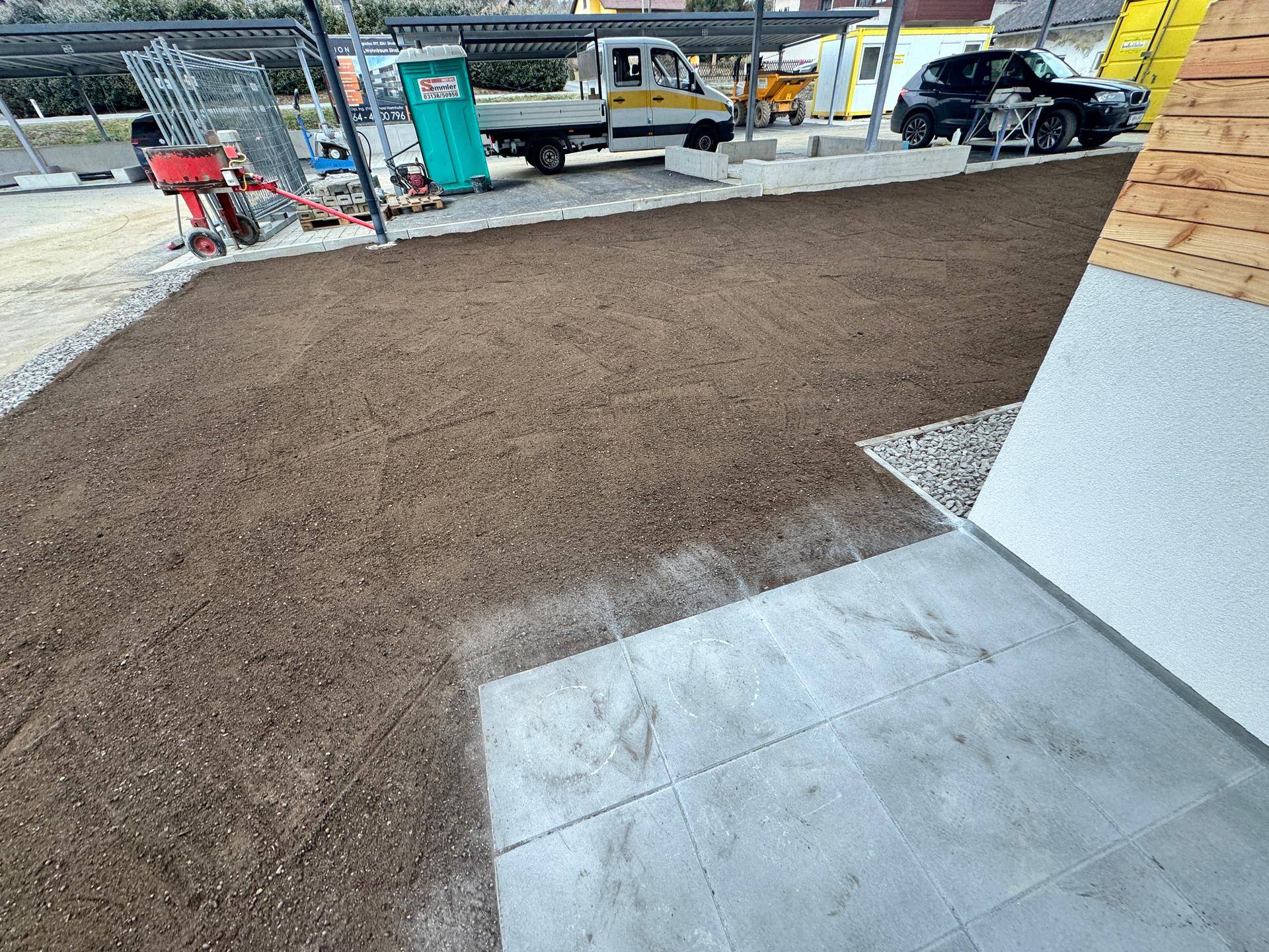 Neu angelegter Gartenbereich mit Erde, bereit für Bepflanzung, angrenzend an einen Parkplatz.