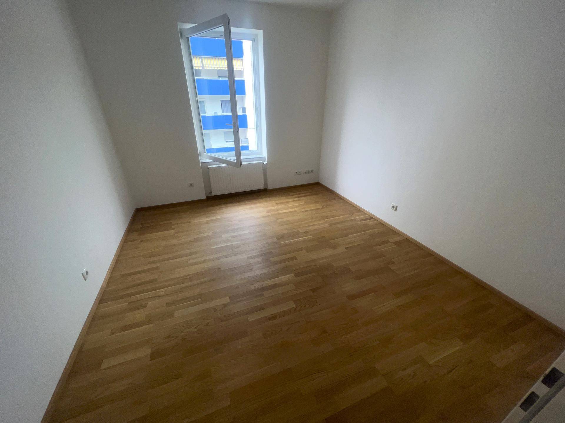 Helles Schlafzimmer mit Parkettboden und einem Fenster mit Blick auf die Umgebung.