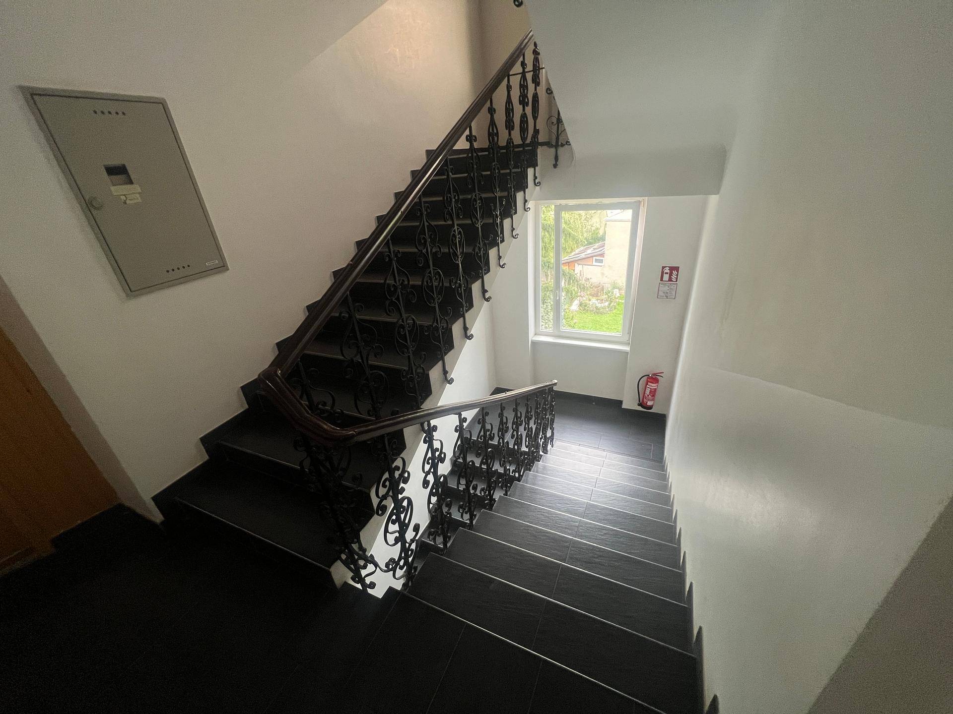 Treppenhaus mit dunklen Fliesen, schmiedeeisernem Geländer und einem Fenster für Tageslicht.