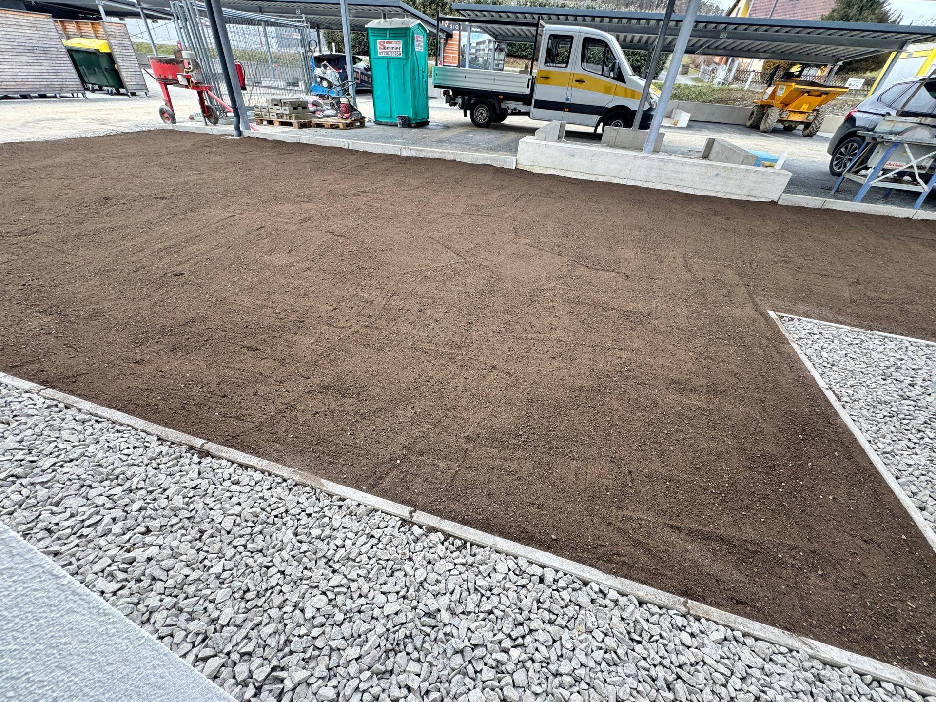 Großer Gartenbereich mit vorbereiteter Erde und Kiesstreifen, umgeben von Parkplätzen.