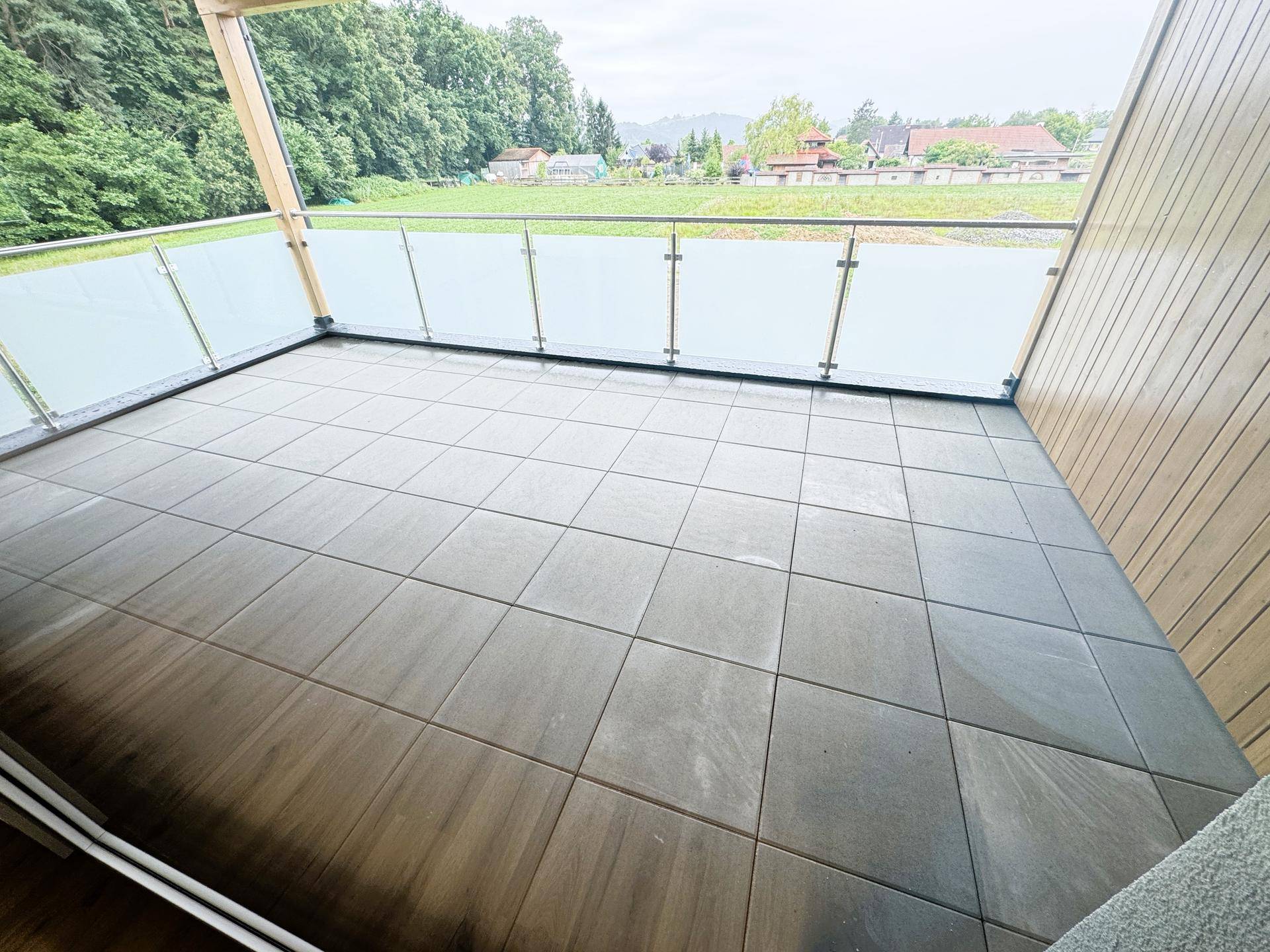 Geräumiger Balkon mit Fliesenboden, Glasgeländer und Blick auf die grüne Umgebung.