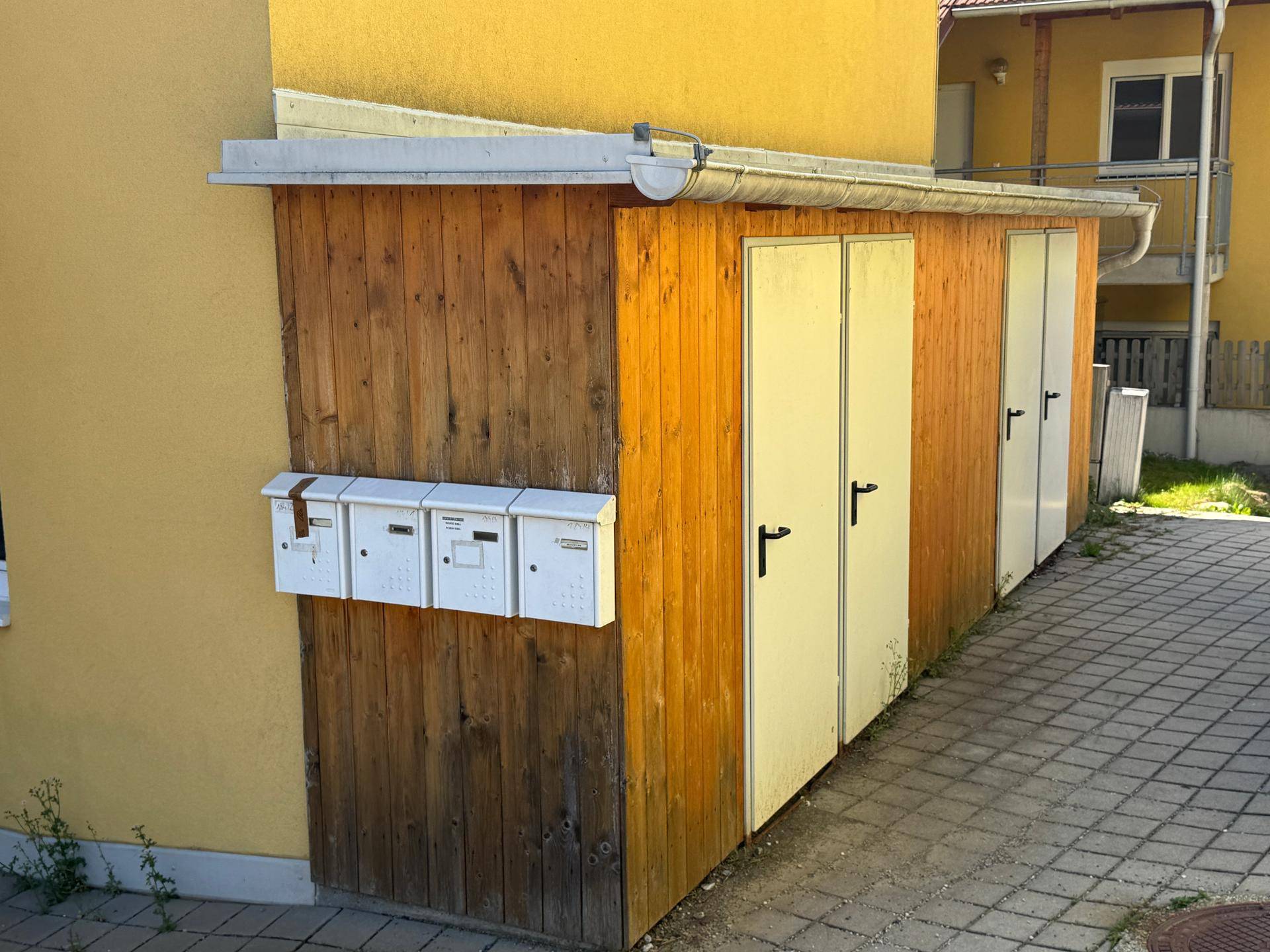 Briefkästen und Holzschuppen an der Seite des gelben Wohngebäudes.