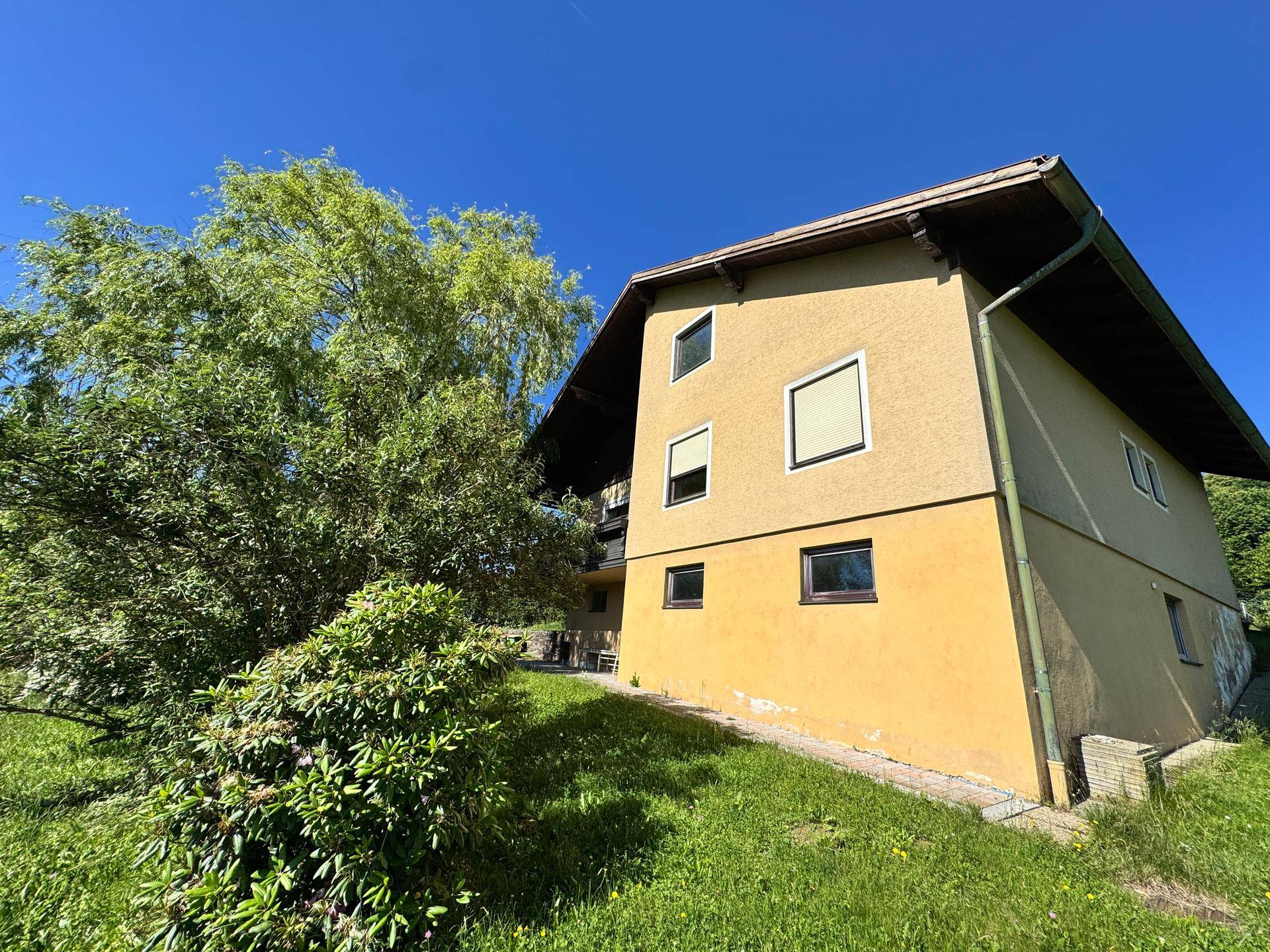 Seitenansicht des Hauses mit gelber Fassade und grüner Umgebung unter blauem Himmel.