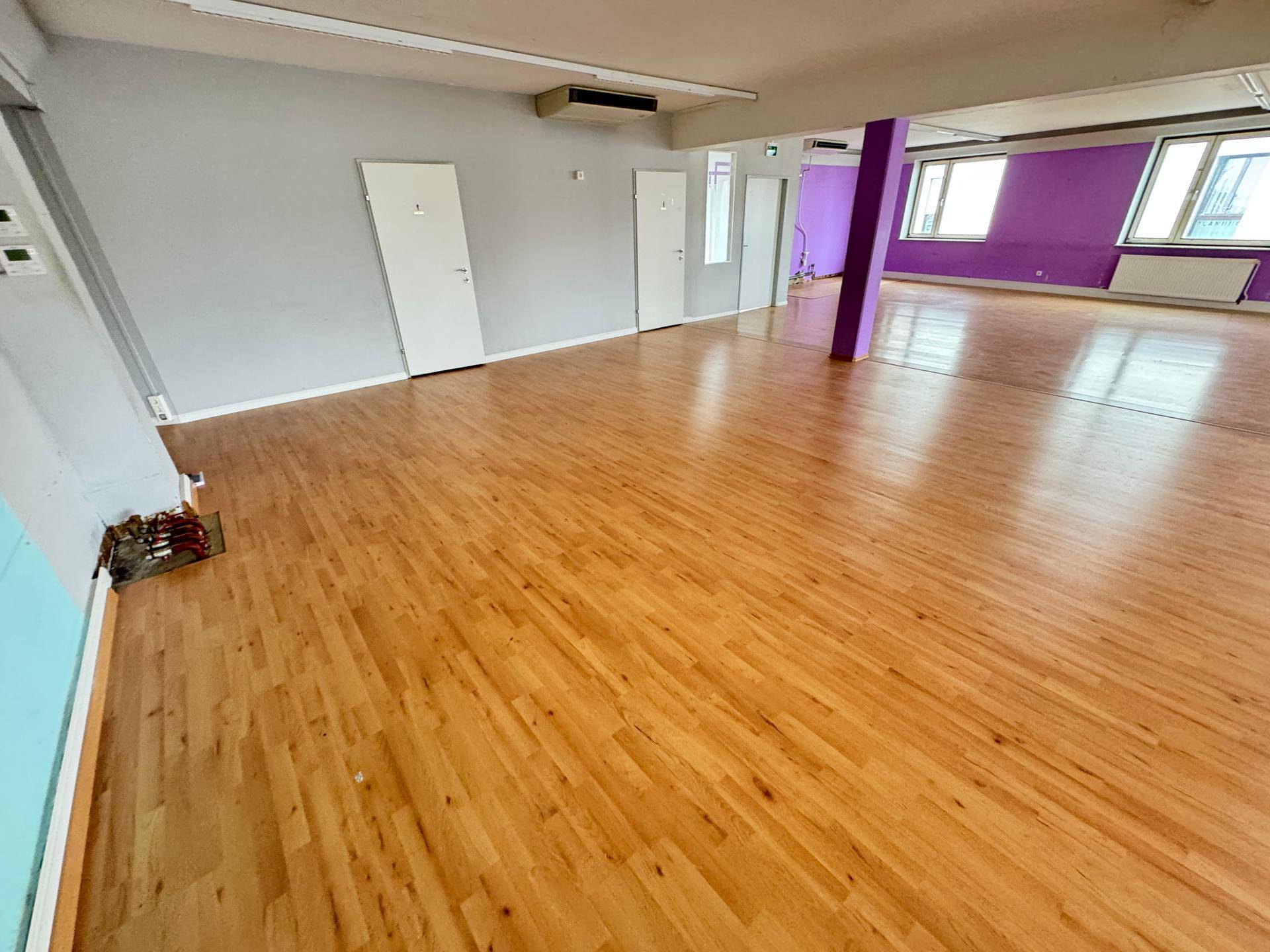 Spacious room with light laminate flooring, several white doors, and a ceiling-mounted air conditioning unit.