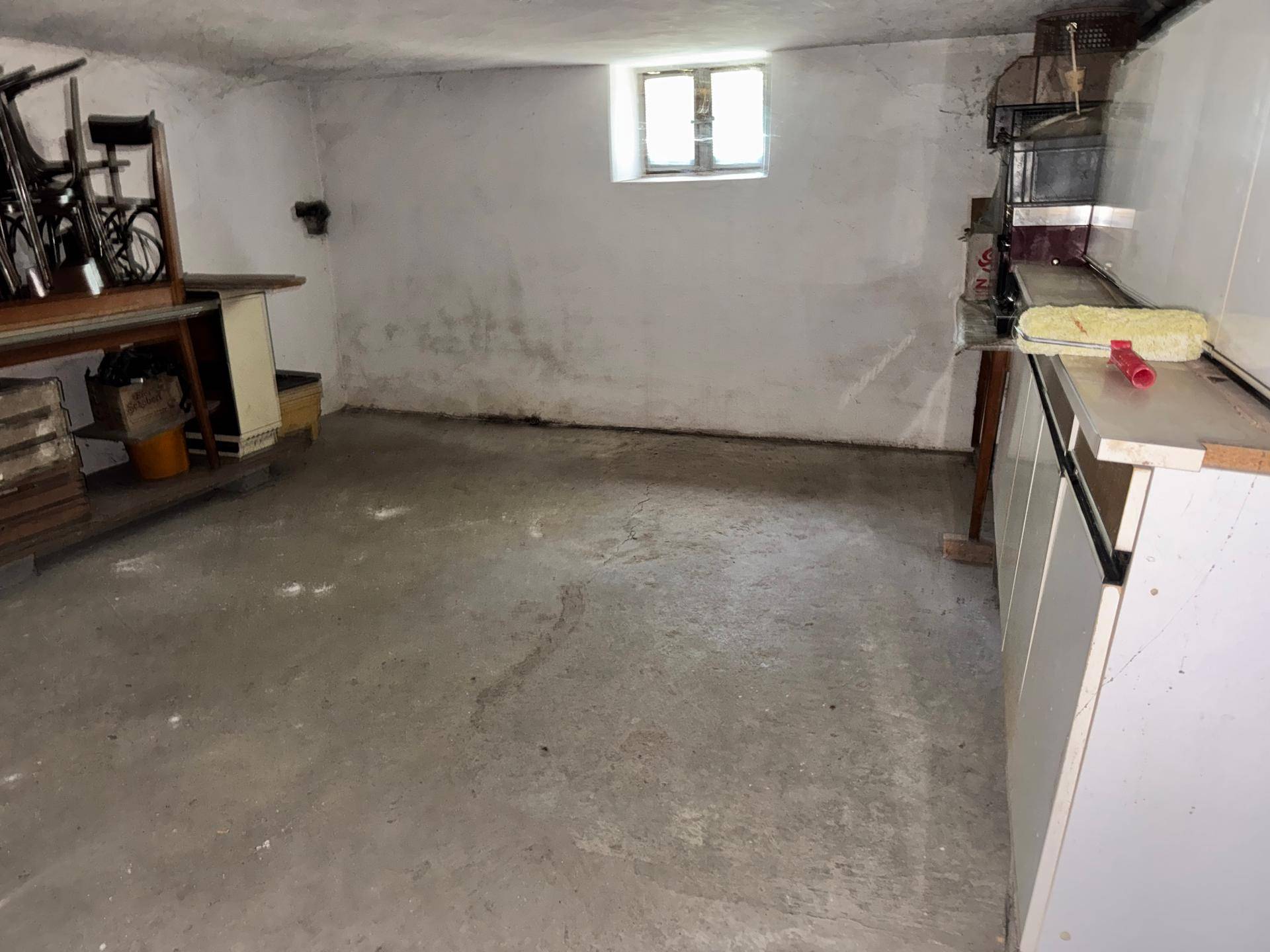 Unfertiger Kellerraum mit Betonboden, kleinem Fenster und Abstellmöglichkeiten für diverse Gegenstände.