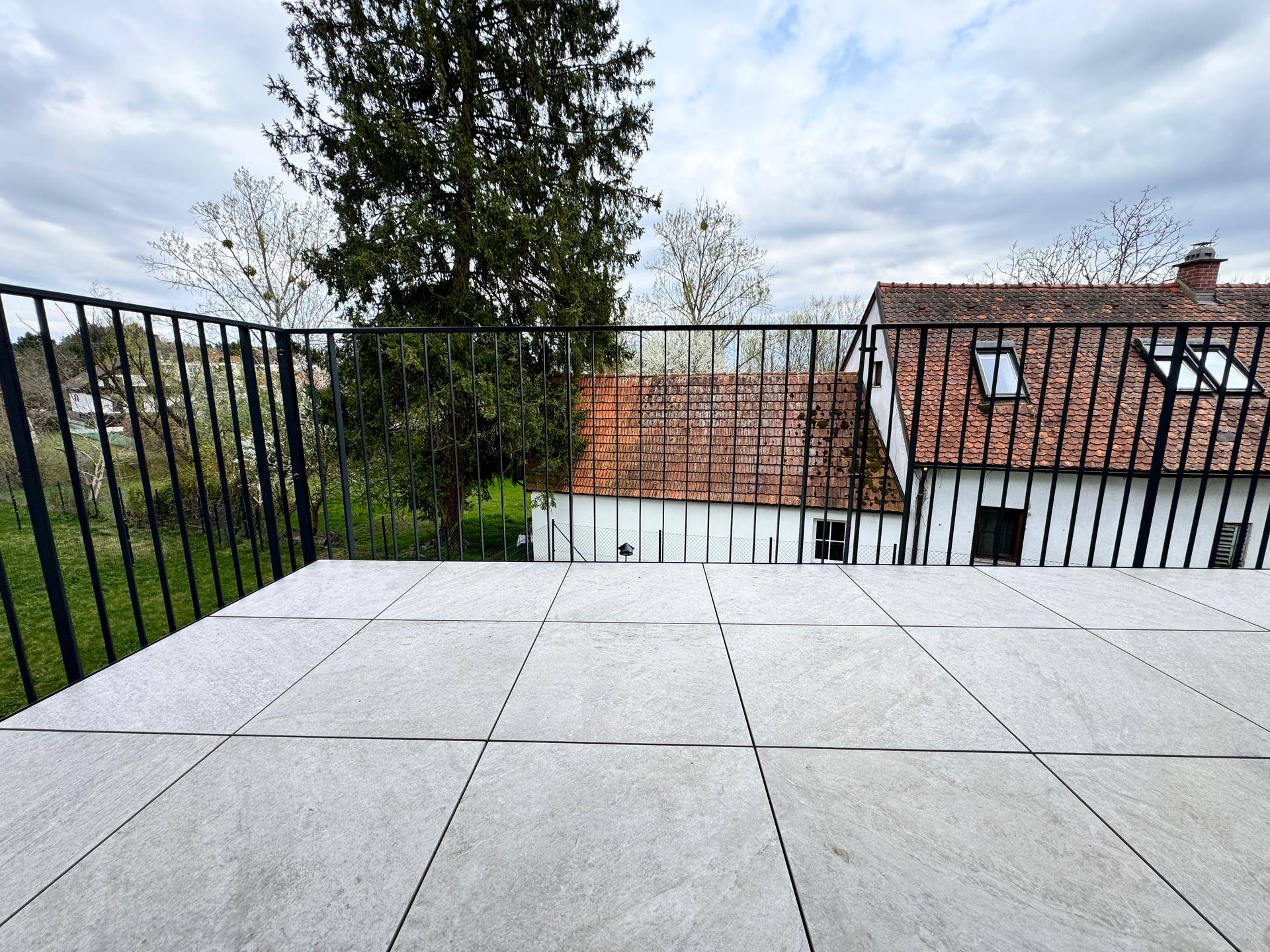 Breiter Balkon mit Fliesenboden und Blick auf Bäume und ein älteres Gebäude.