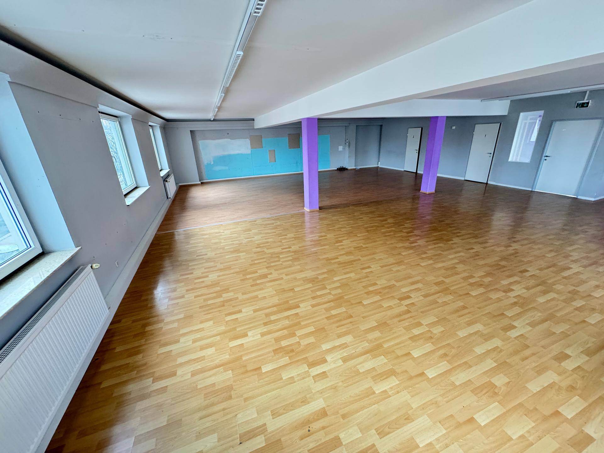Weitläufiger Raum mit hellem Laminatboden, verschiedenen Wandfarben und vielen Fenstern.