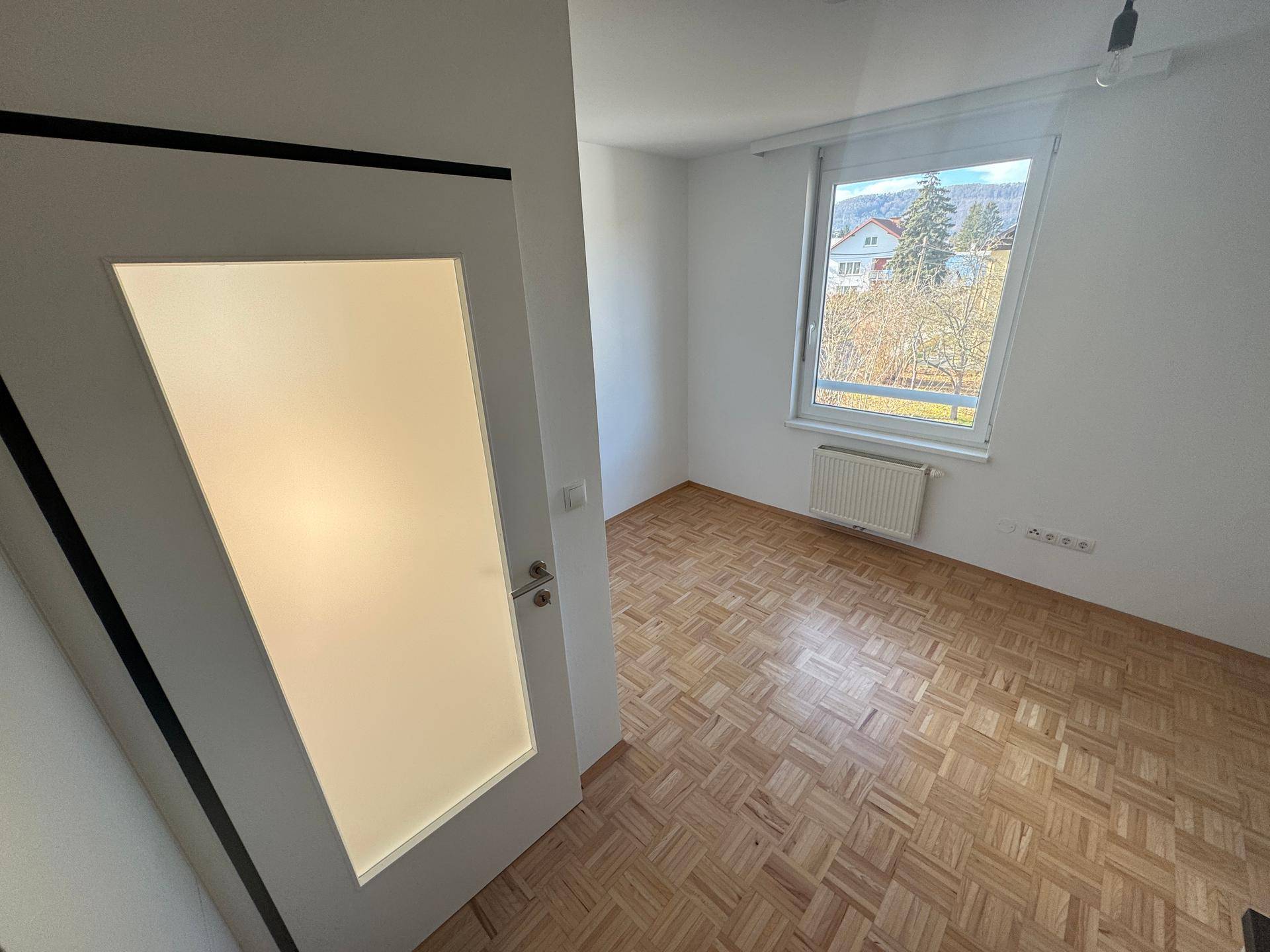 Heller Raum mit Parkettboden, großem Fenster und Blick ins Grüne.
