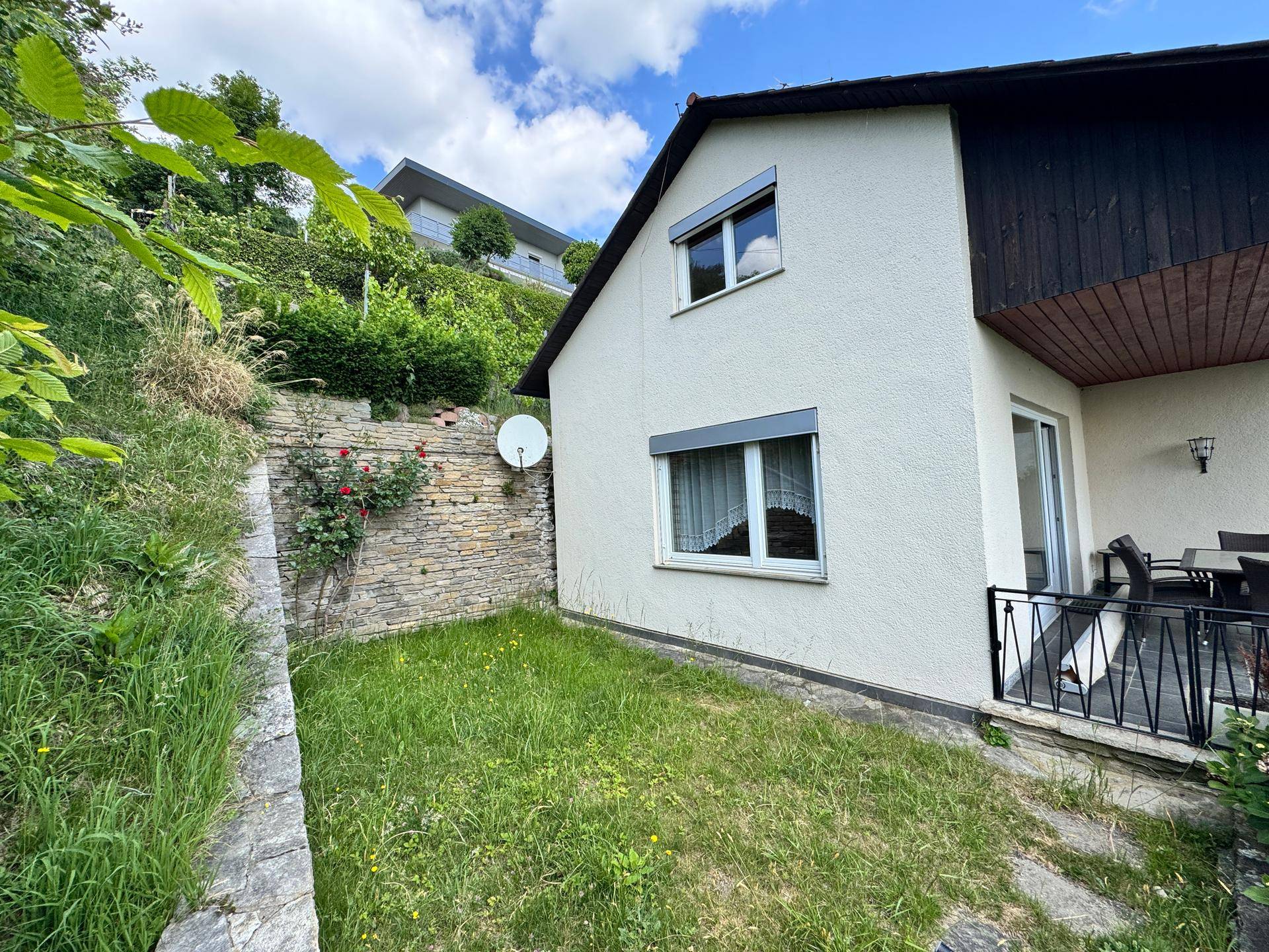 Ansicht des Hauses von unten mit kleiner Rasenfläche und Balkon, umgeben von grünen Büschen.