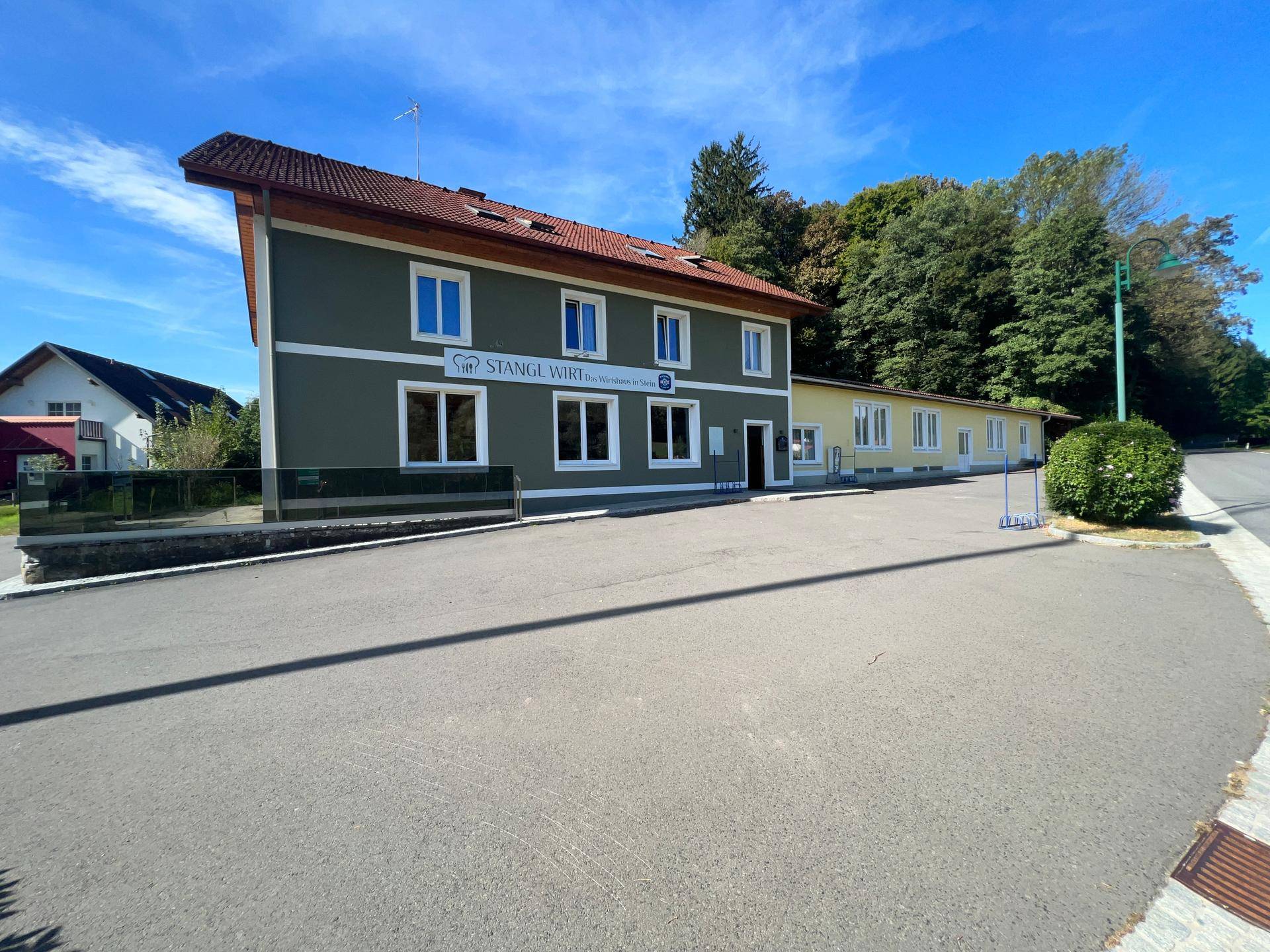 Vorderansicht des Gebäudes mit großem asphaltierten Parkplatz und dem Schriftzug "Stangl Wirt".