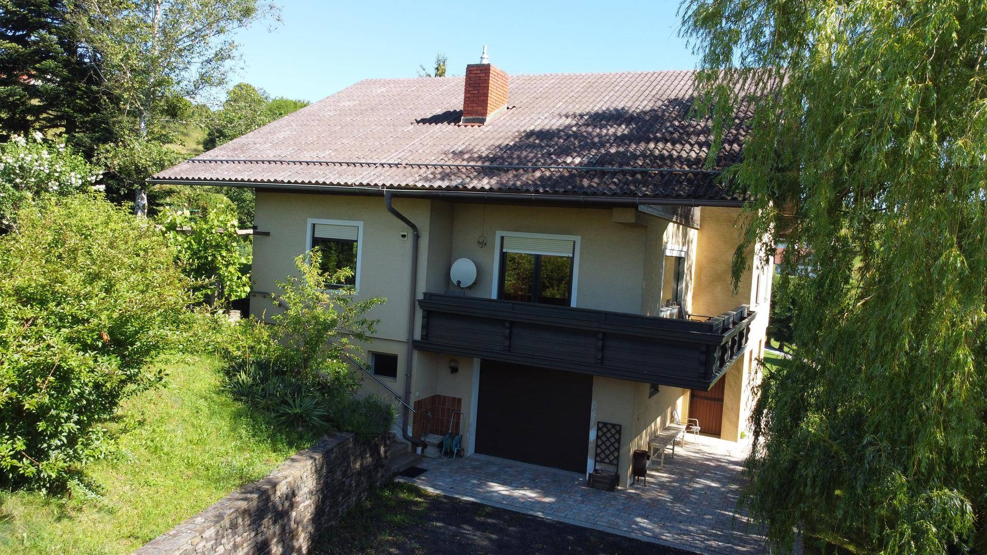 Haus mit Satteldach und Balkon, umgeben von viel Grün und Bäumen.
