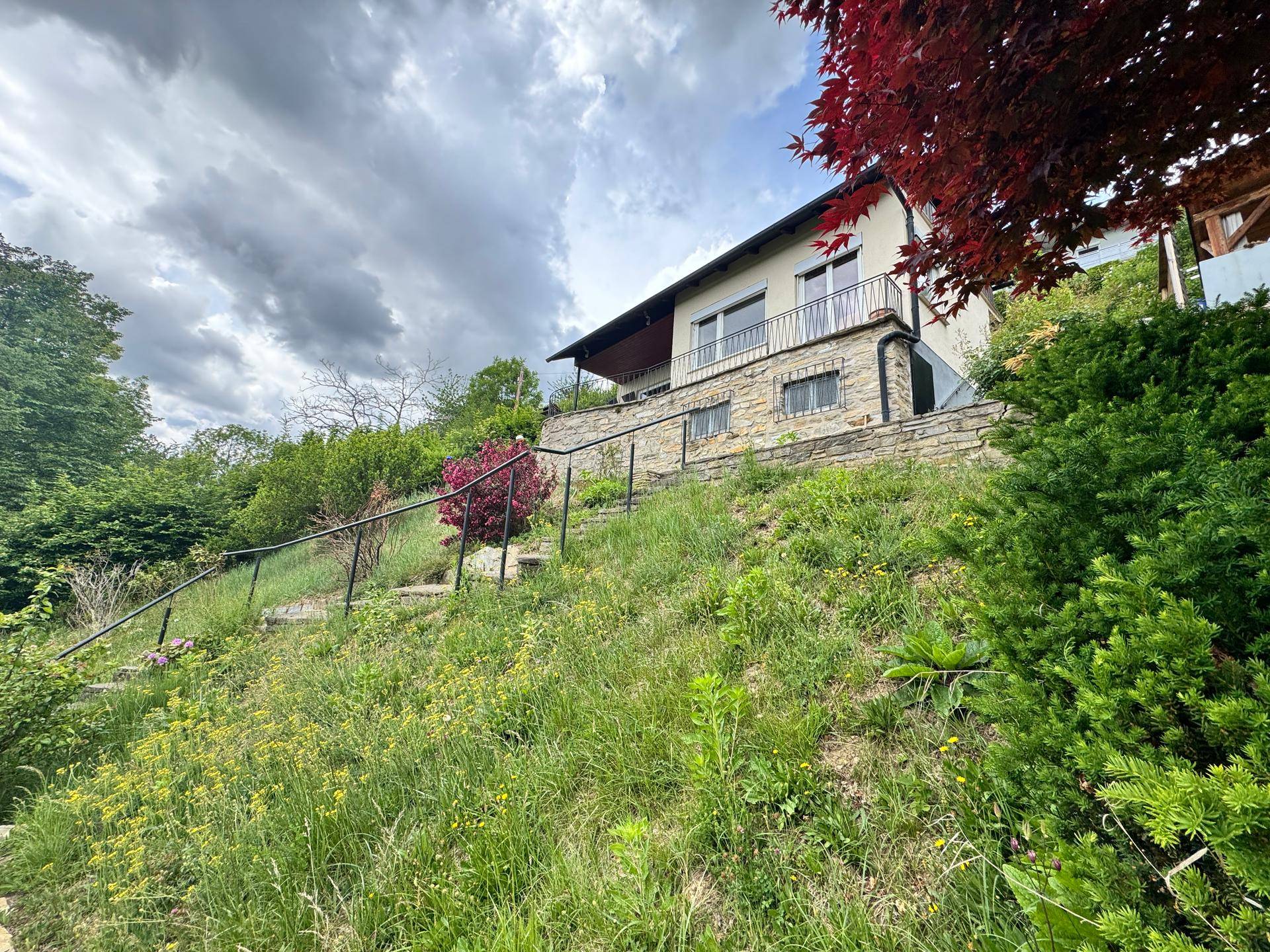 Haus auf einem Hanggrundstück mit Steinterrassen und üppiger Vegetation unter dramatischem Himmel.