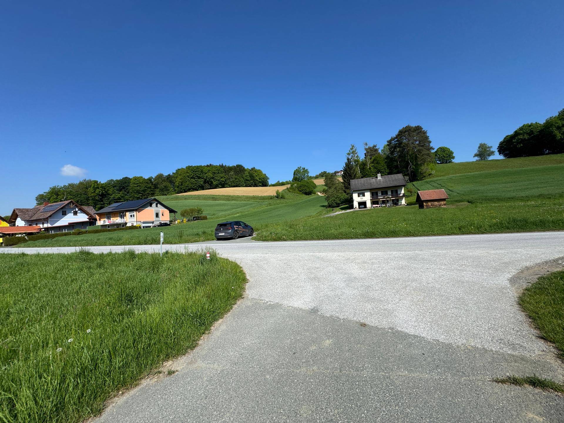 Ländliche Umgebung mit grünen Hügeln, vereinzelten Häusern und einer Straße unter blauem Himmel.