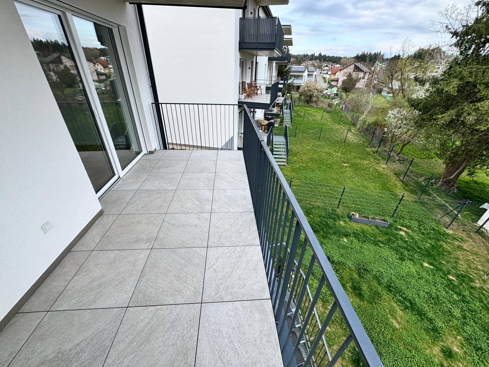 Langer Balkon mit Fliesenboden und Metallgeländer, bietet Blick auf die grüne Umgebung.
