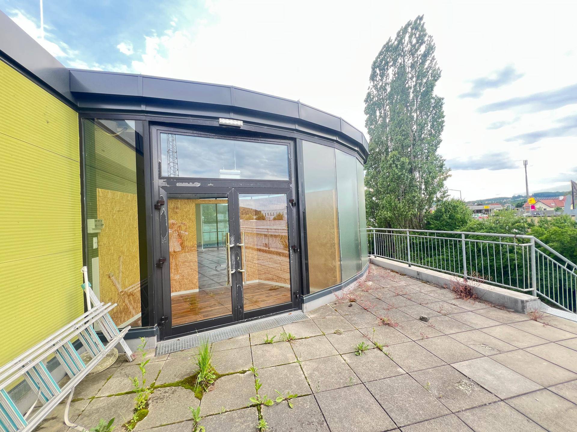 Terrasse mit grauen Fliesen und Metallgeländer, angrenzend an die Glasfassade des Gebäudes mit Baumblick.