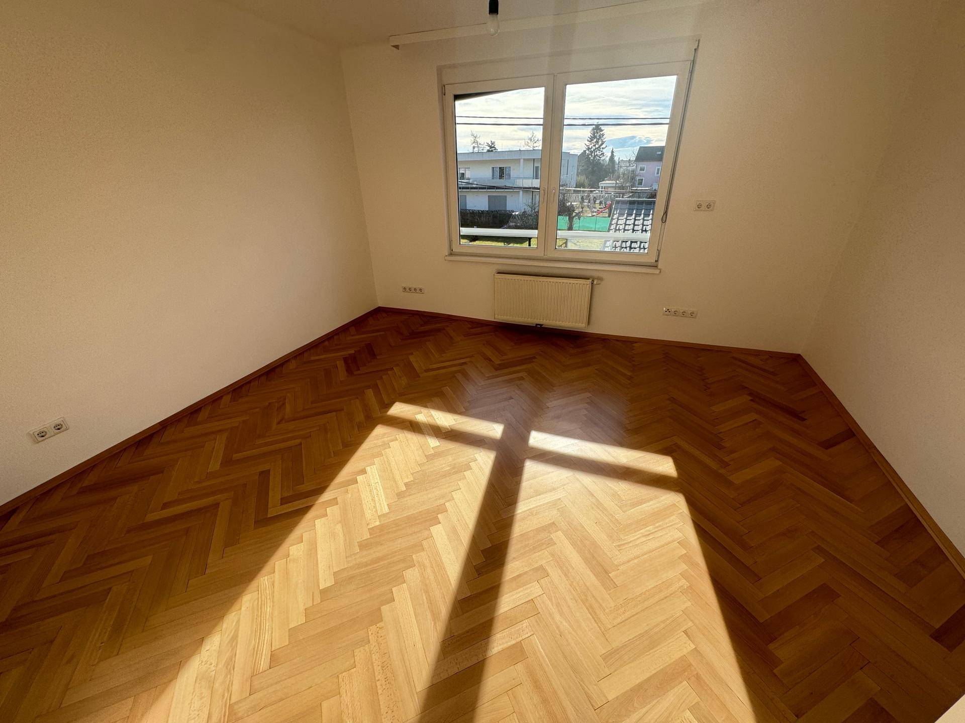 Heller Raum mit Parkettboden und großem Fenster mit Sonneneinstrahlung.