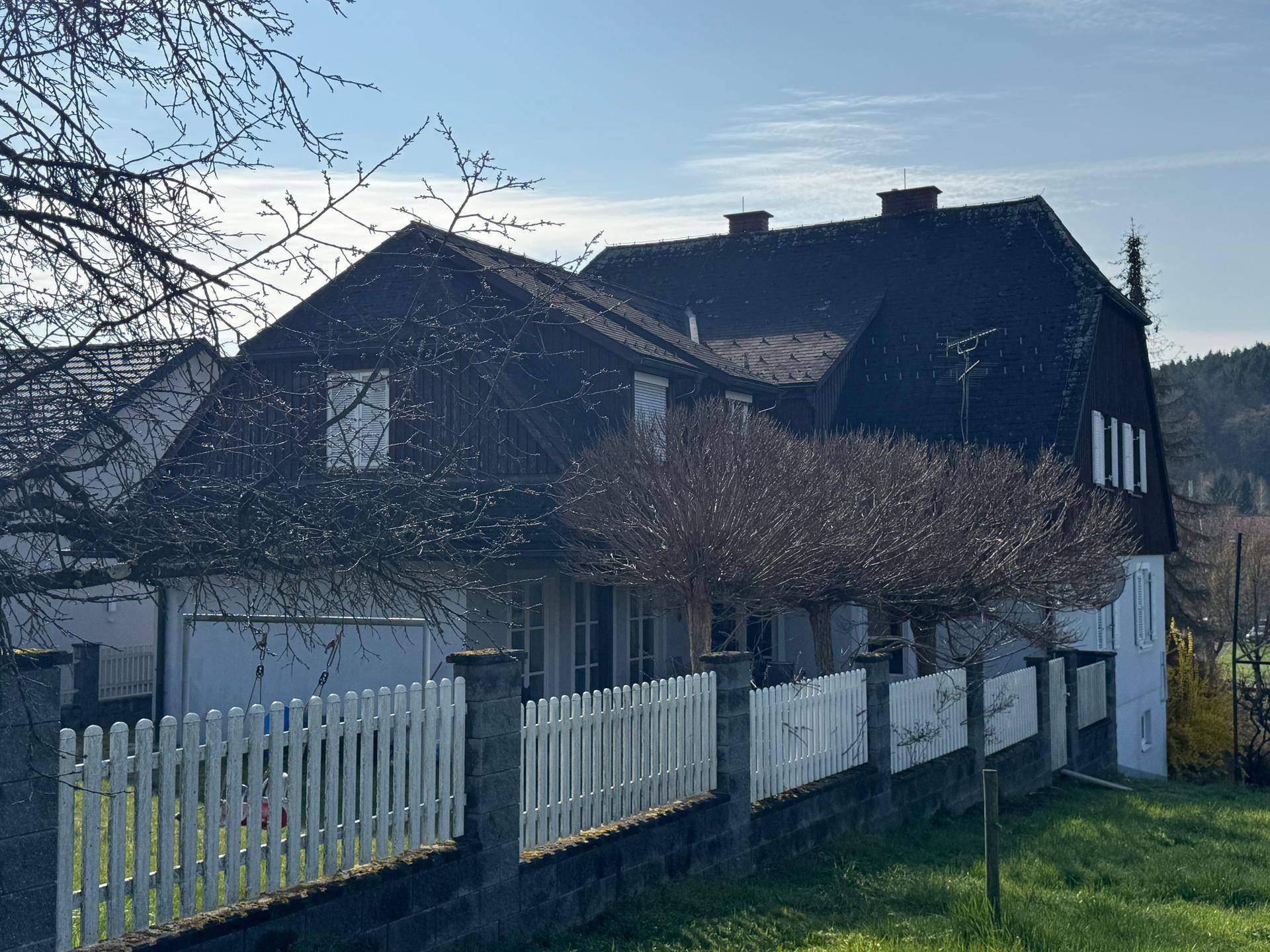 Rückseite des Hauses mit Mansarddach, weißem Zaun und Blick auf die umliegende Natur.