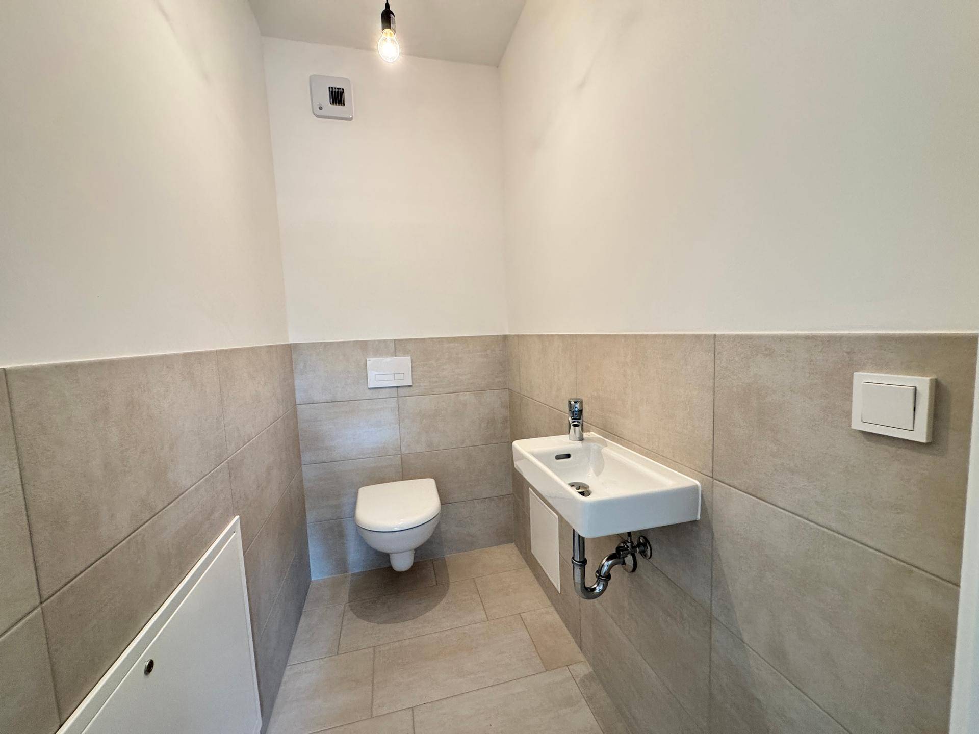 Modernes Gäste-WC mit wandhängender Toilette, kleinem Waschbecken und beigefarbenen Fliesen.