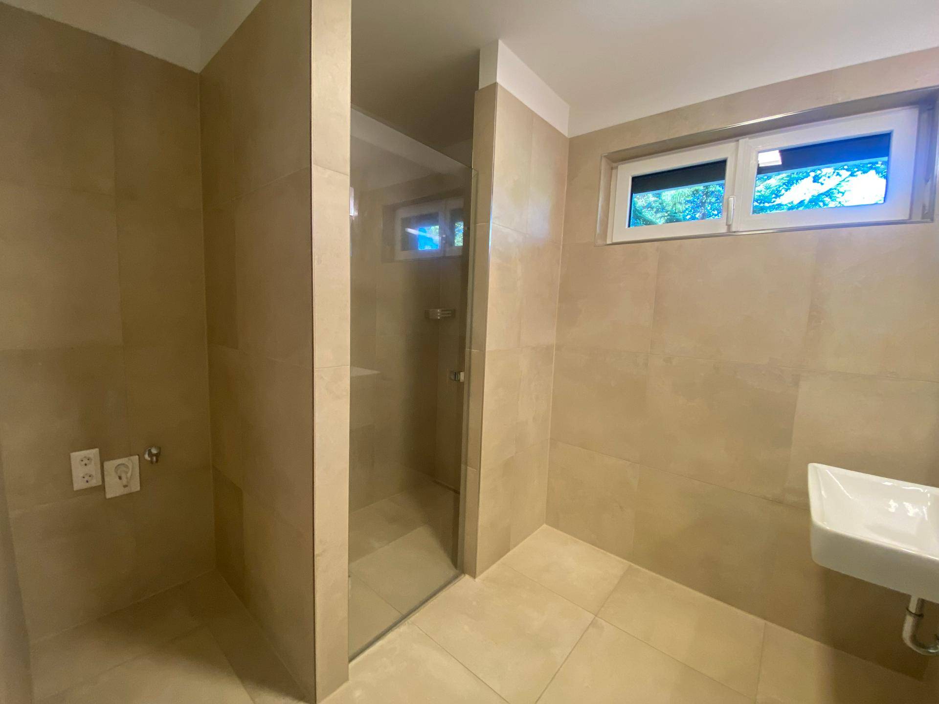 Modernes Badezimmer mit beigefarbenen Fliesen, ebenerdiger Dusche und Fenster für Tageslicht.